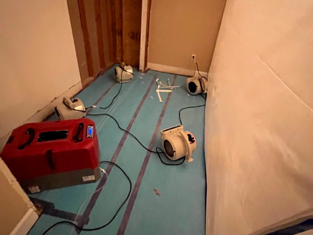Room under renovation with construction equipment, including dehumidifier, some air movers, inside a containment for dust control, on a blue protective floor covering.