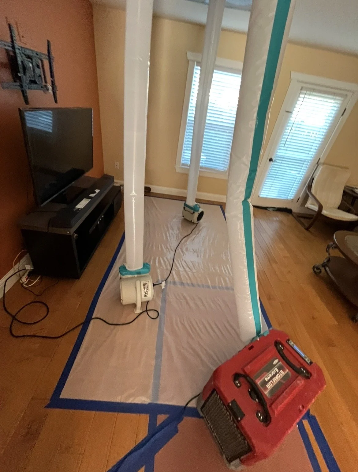 Room with two large vertical poles, covered in plastic, set up on a plastic sheet taped to hardwood floor, with a fan near the poles. There is a TV on a stand against an orange wall, a chair near a glass door with blinds, and a red toolbox on the floor.