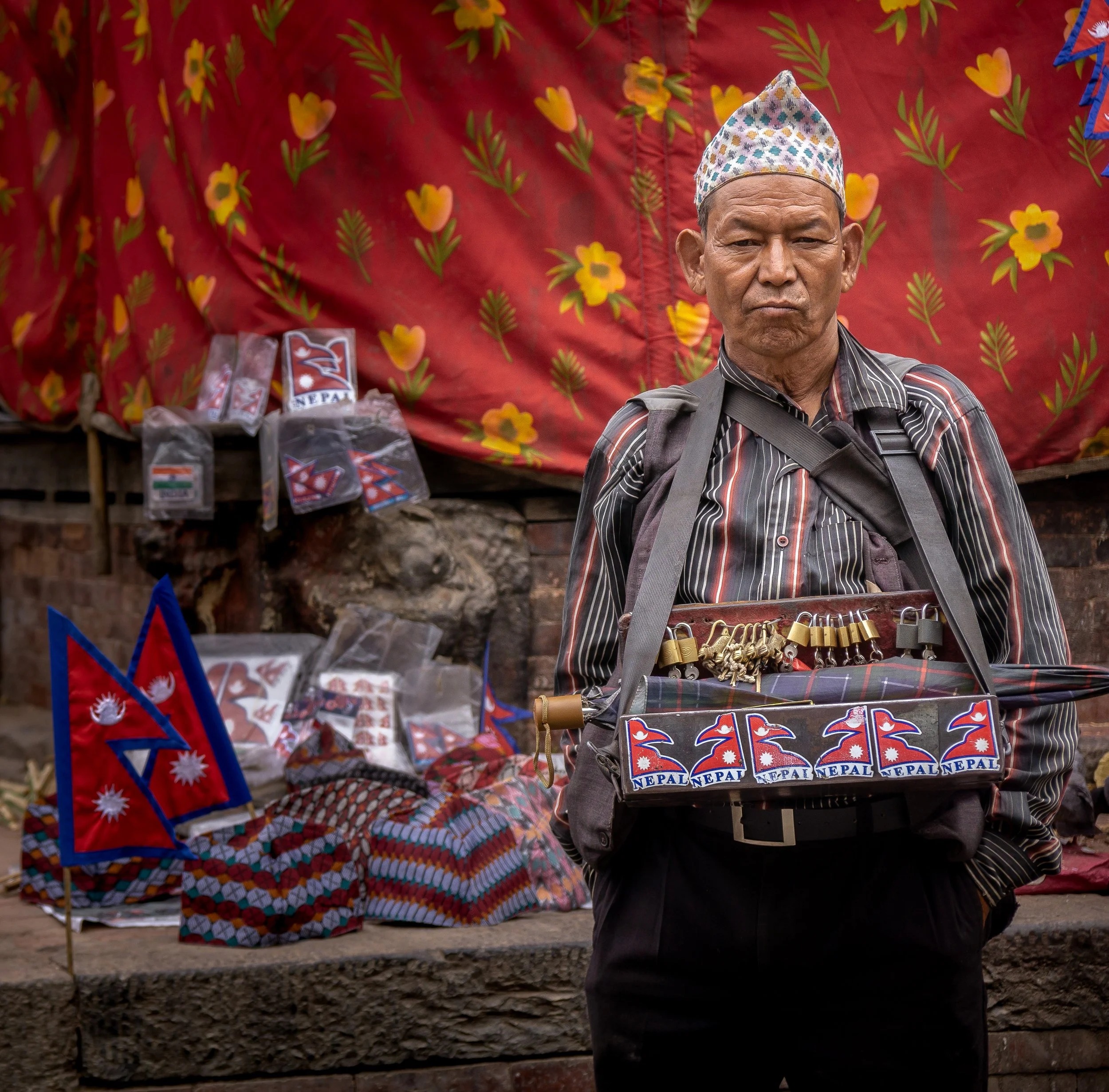 An older man with a serious expression wearing a patterned hat and striped shirt, standing in front of a colorful banner and Nepali flags, with small packages of items on display.