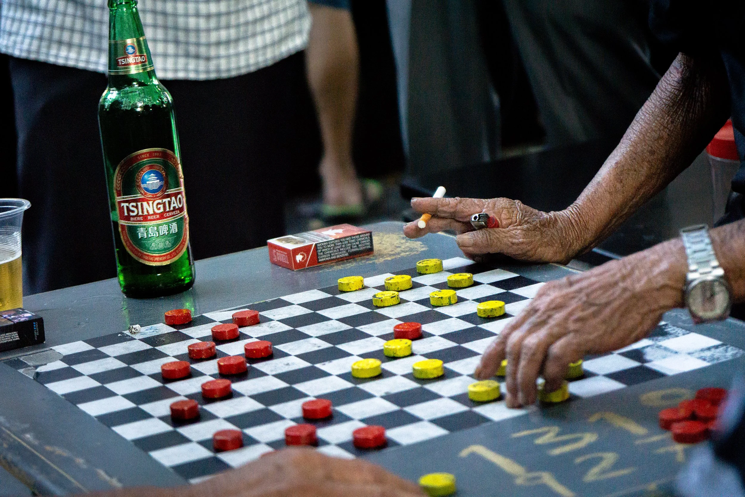 Close-up of people playing a game of checkers on a black-and-white checkered board, with yellow and red game pieces, with a green Tsingtao beer bottle and miscellaneous objects on the table.