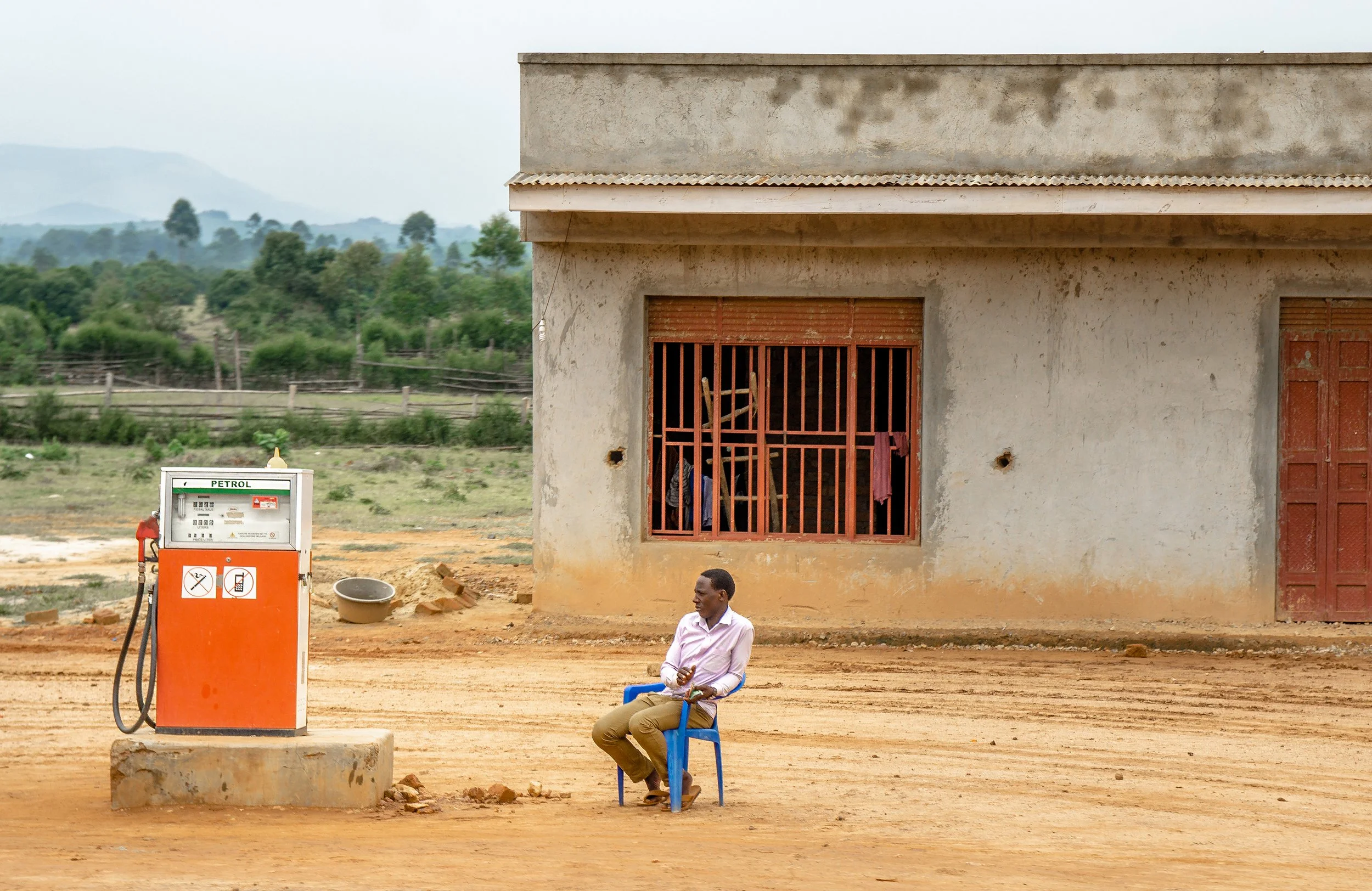 A man sitting on a blue plastic chair at a fuel station with a petrol pump in the foreground. A building with two windows and a door is in the background, along with green trees and mountains.