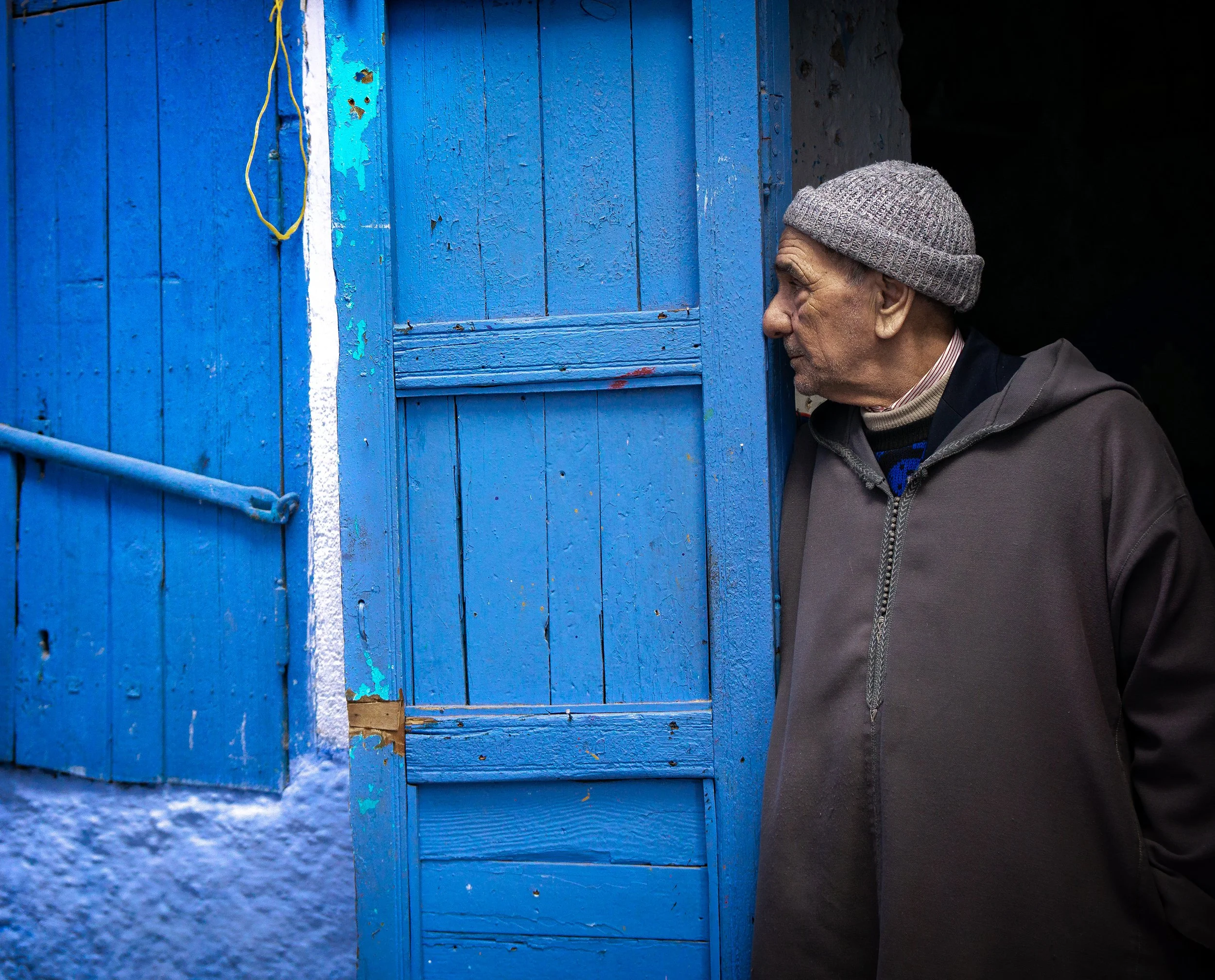 A man wearing a gray knit beanie, a dark jacket, and a collared shirt peeks into a blue wooden door with peeling paint and a weathered appearance, set against a dark background.