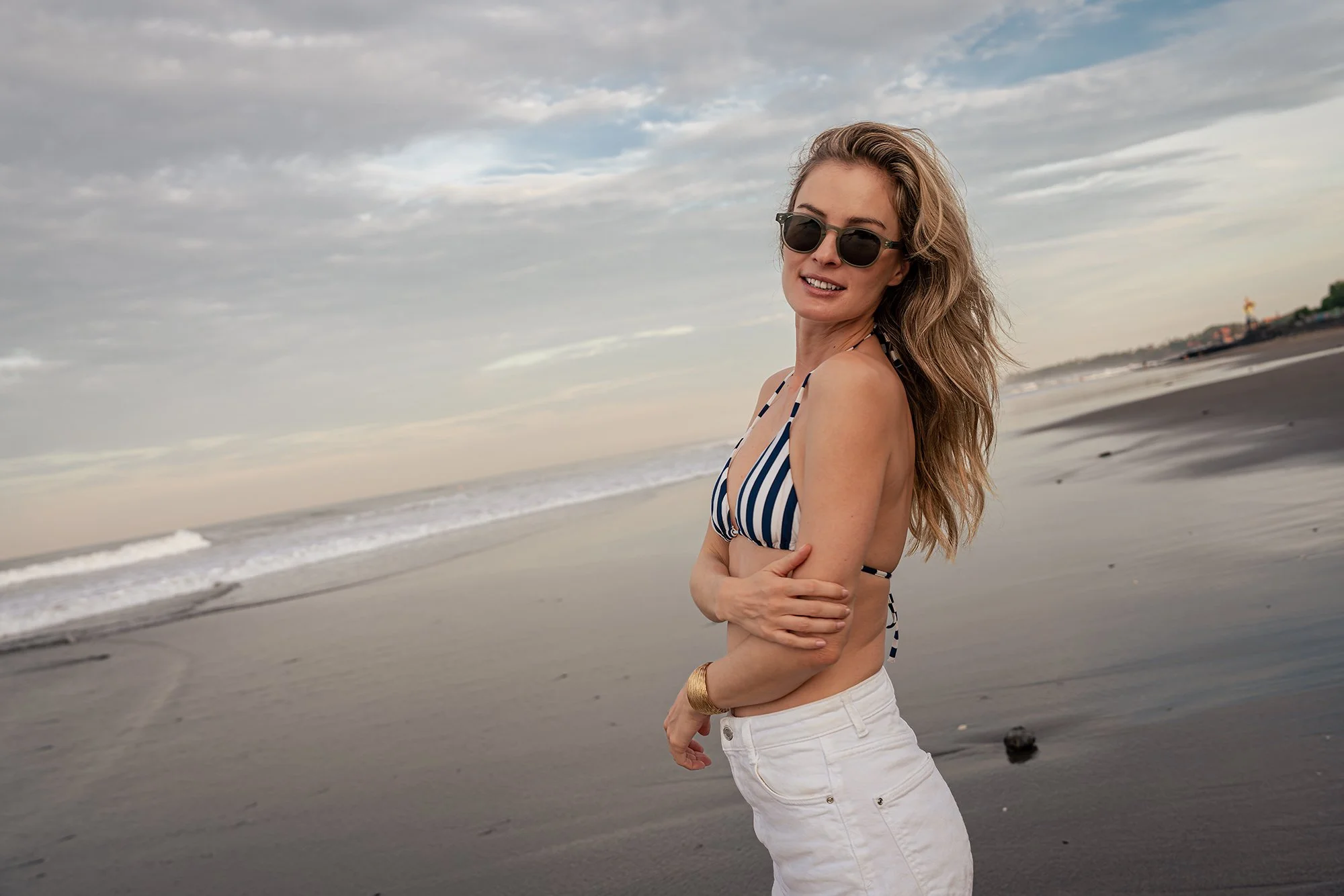 A woman with wavy blonde hair wearing sunglasses, a striped bikini top, and white pants stands on a beach with her arms crossed, looking at the camera with a smile as waves and a cloudy sky are in the background.