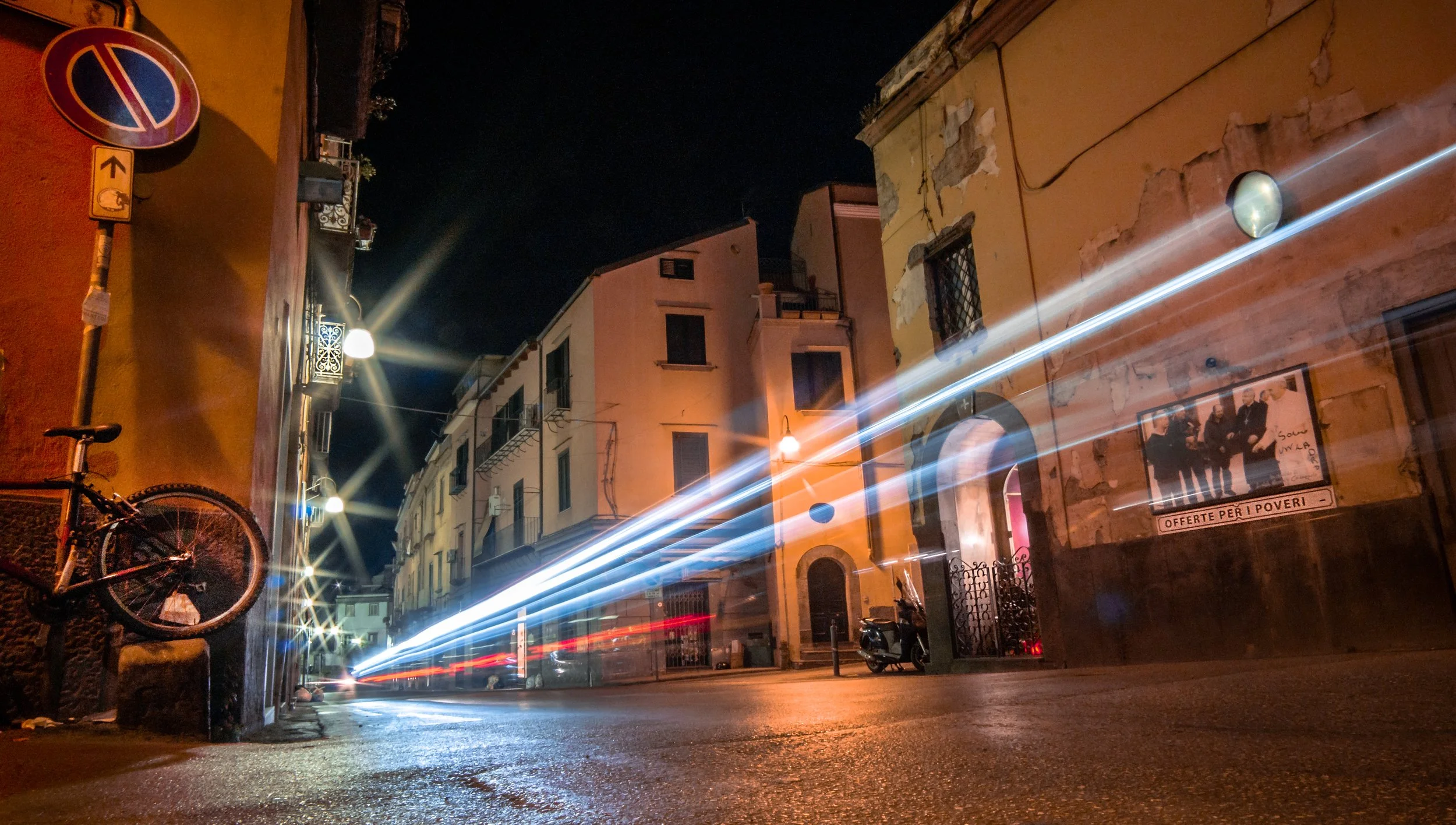 Nighttime street scene with illuminated buildings, a bicycle mounted on a wall, and streaks of light from moving vehicles, capturing light trails.