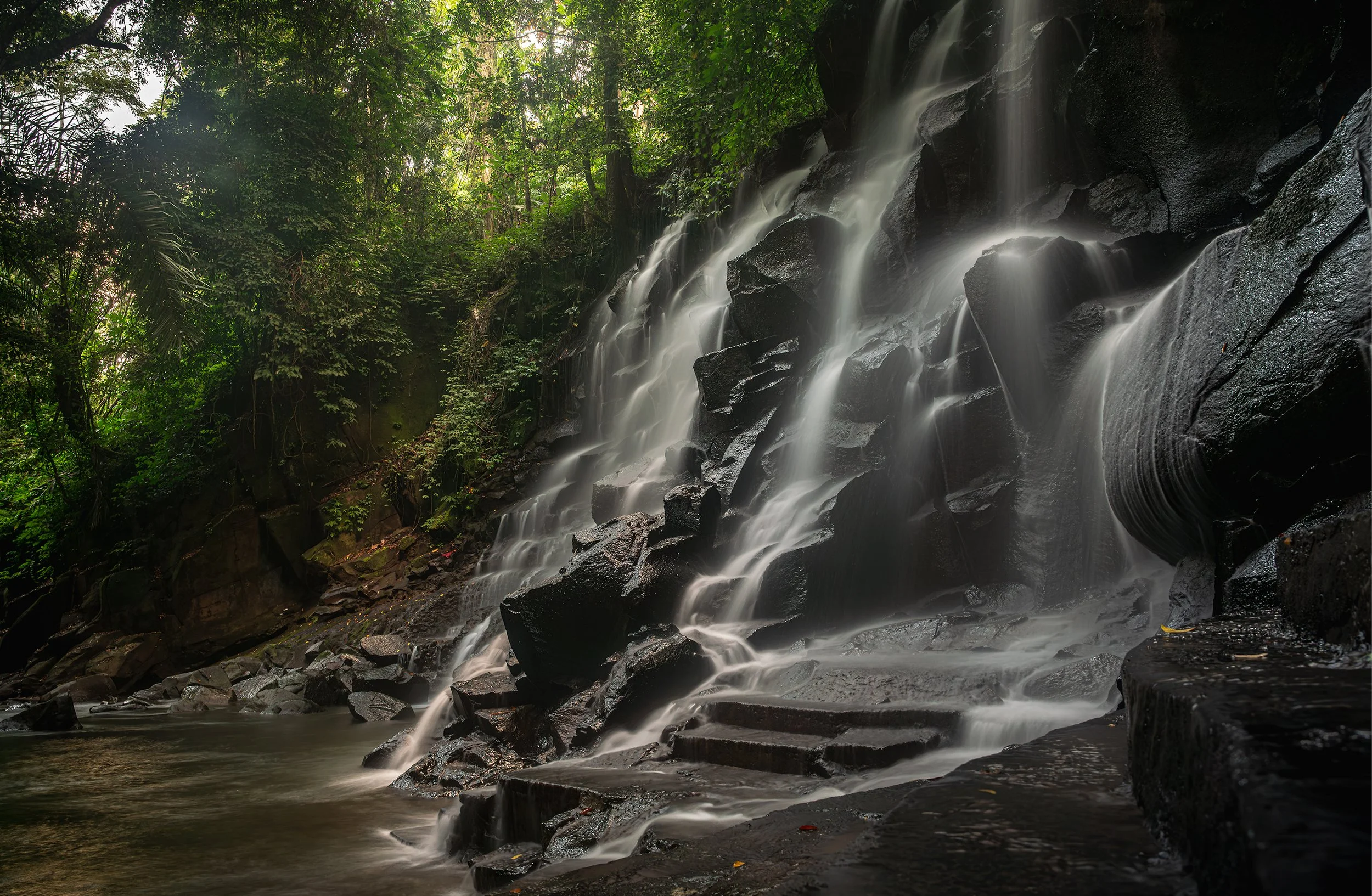 A waterfall flowing over rocks in a lush green forest with sunlight streaming through the trees.