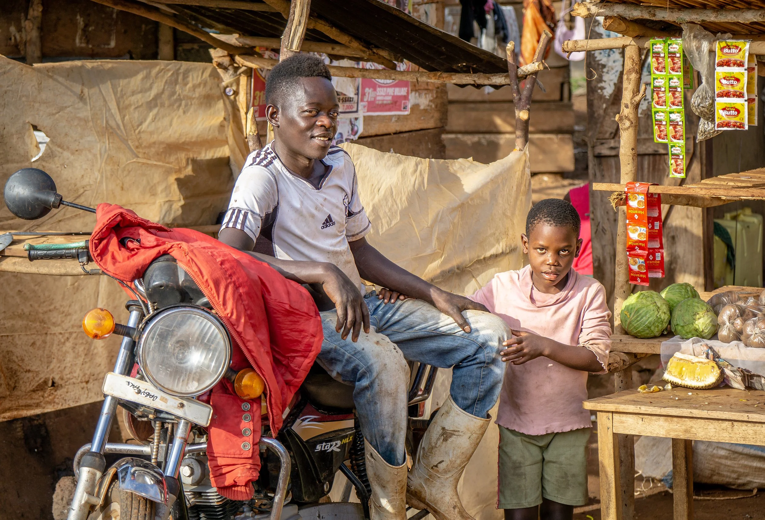 A man and a boy standing at a small outdoor market stall with produce and packaged goods, with the man sitting on a motorcycle.