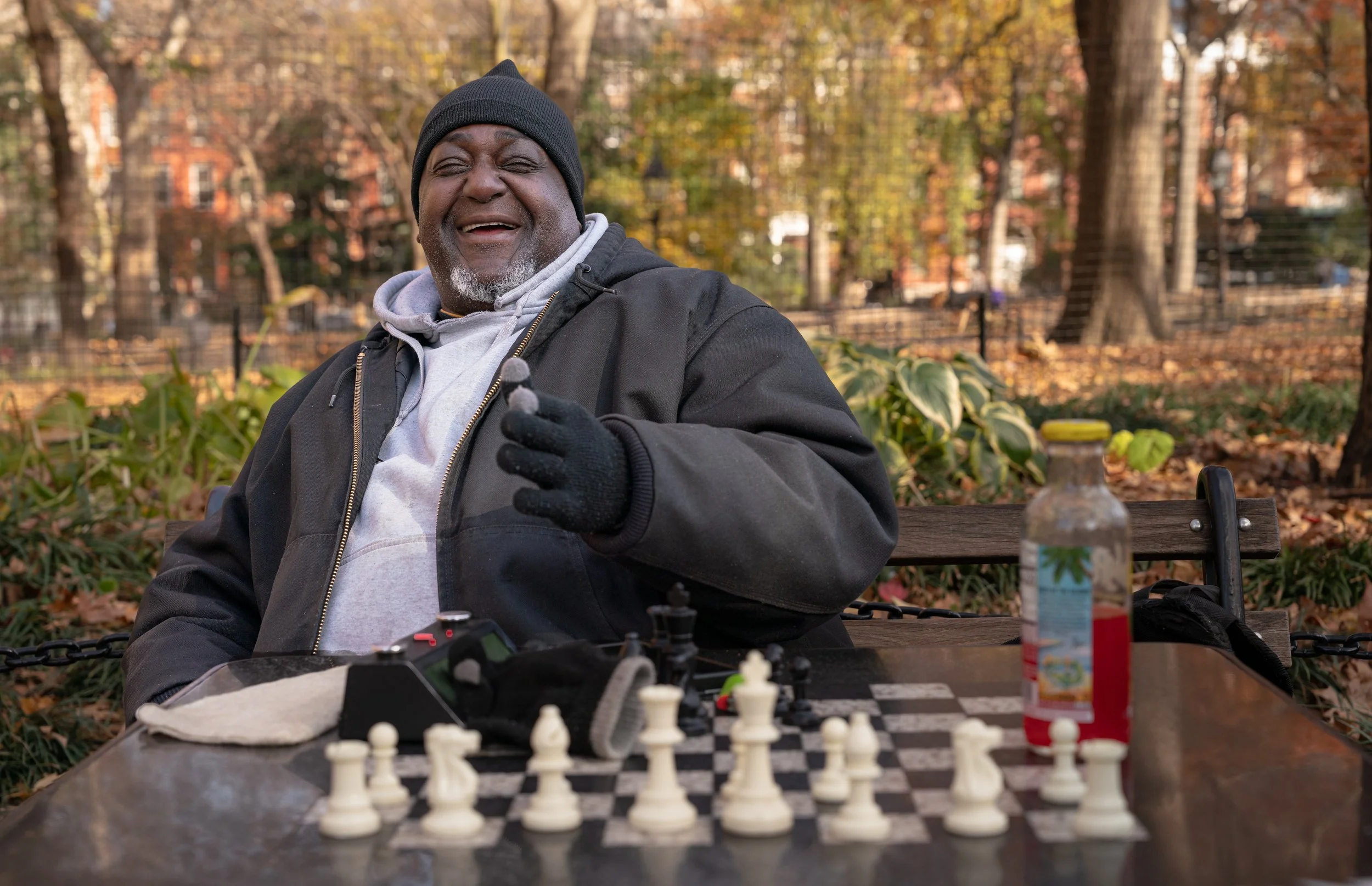 An elderly man with dark skin, gray beard, and wearing a black beanie, black jacket, and gray hoodie, laughs and smiles while sitting at a park bench during autumn. In front of him is a chess board with chess pieces, and a water bottle is placed on t