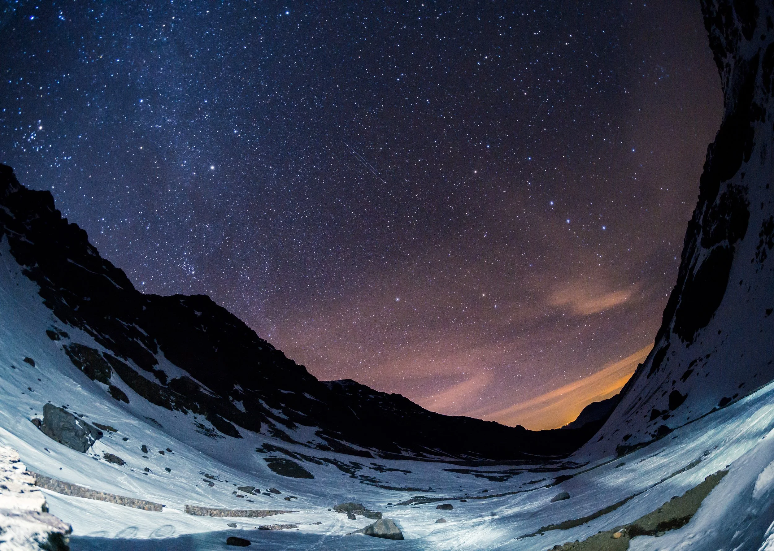 Snow-covered mountain valley under a starry night sky with visible stars and a faint trail of shooting stars