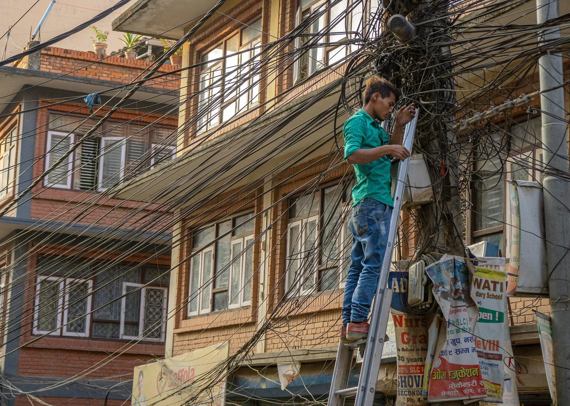 A man standing on a ladder fixing or managing a tangled mass of electrical wires on a utility pole in front of a building with brick and glass windows.