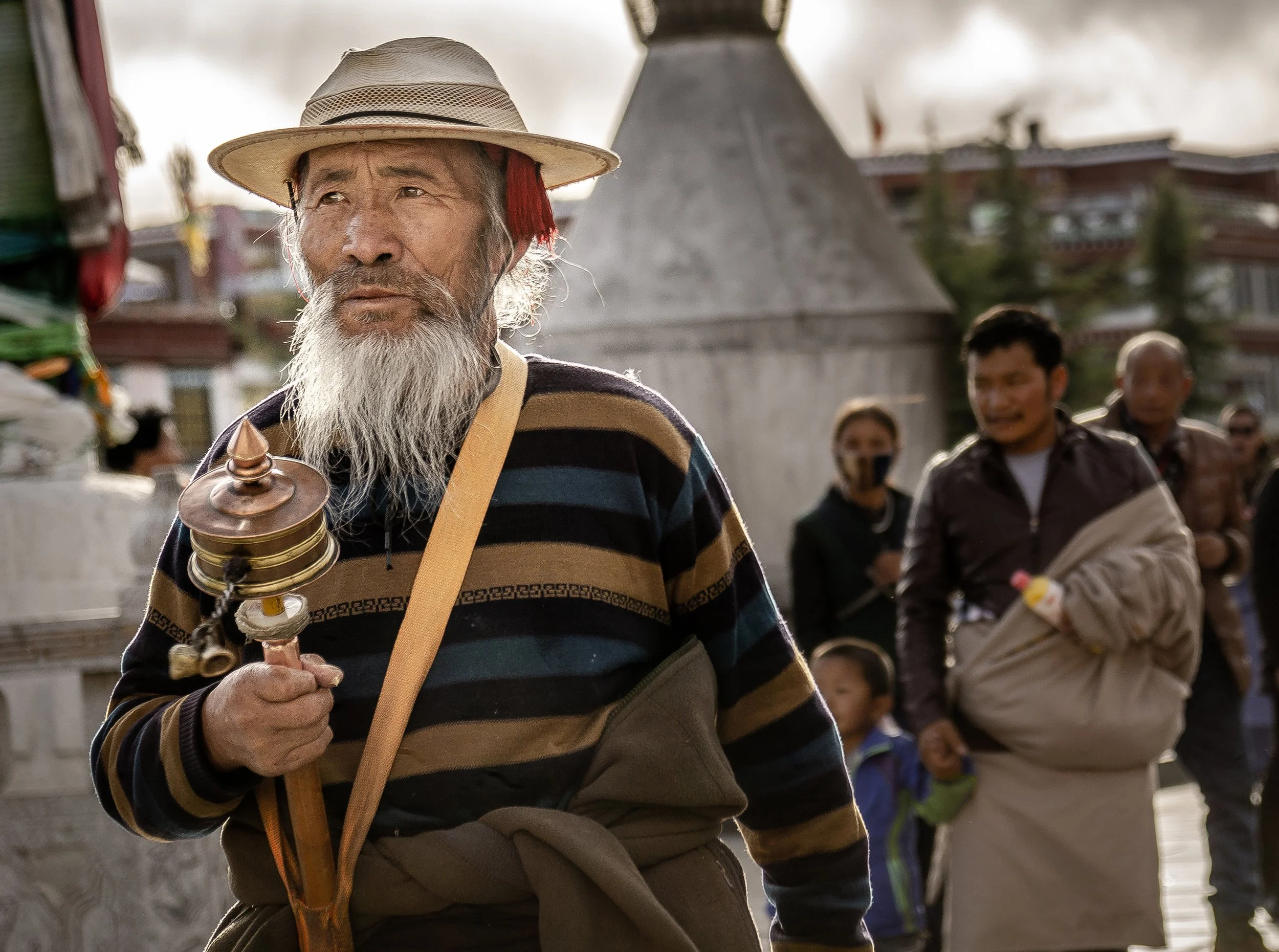 An elderly man with a white beard holding a traditional musical instrument, wearing a striped sweater and a hat, outdoors with a group of people and a religious monument in the background.