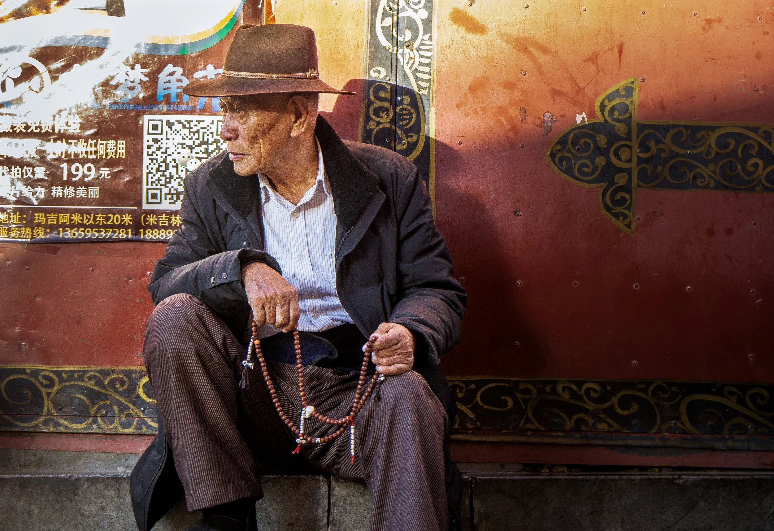 An elderly man wearing a wide-brimmed hat, a dark jacket, and striped pants sitting against a colorful, weathered wall with Chinese characters, QR codes, and decorative patterns, holding prayer beads.