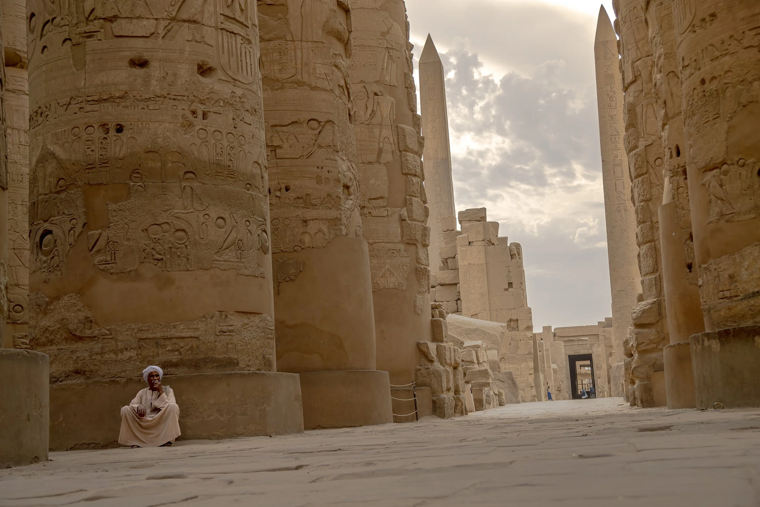 Ancient Egyptian temple ruin with large stone columns covered in hieroglyphics, and a man sitting on the ground in traditional clothing, looking at the camera, with a cloudy sky in the background.