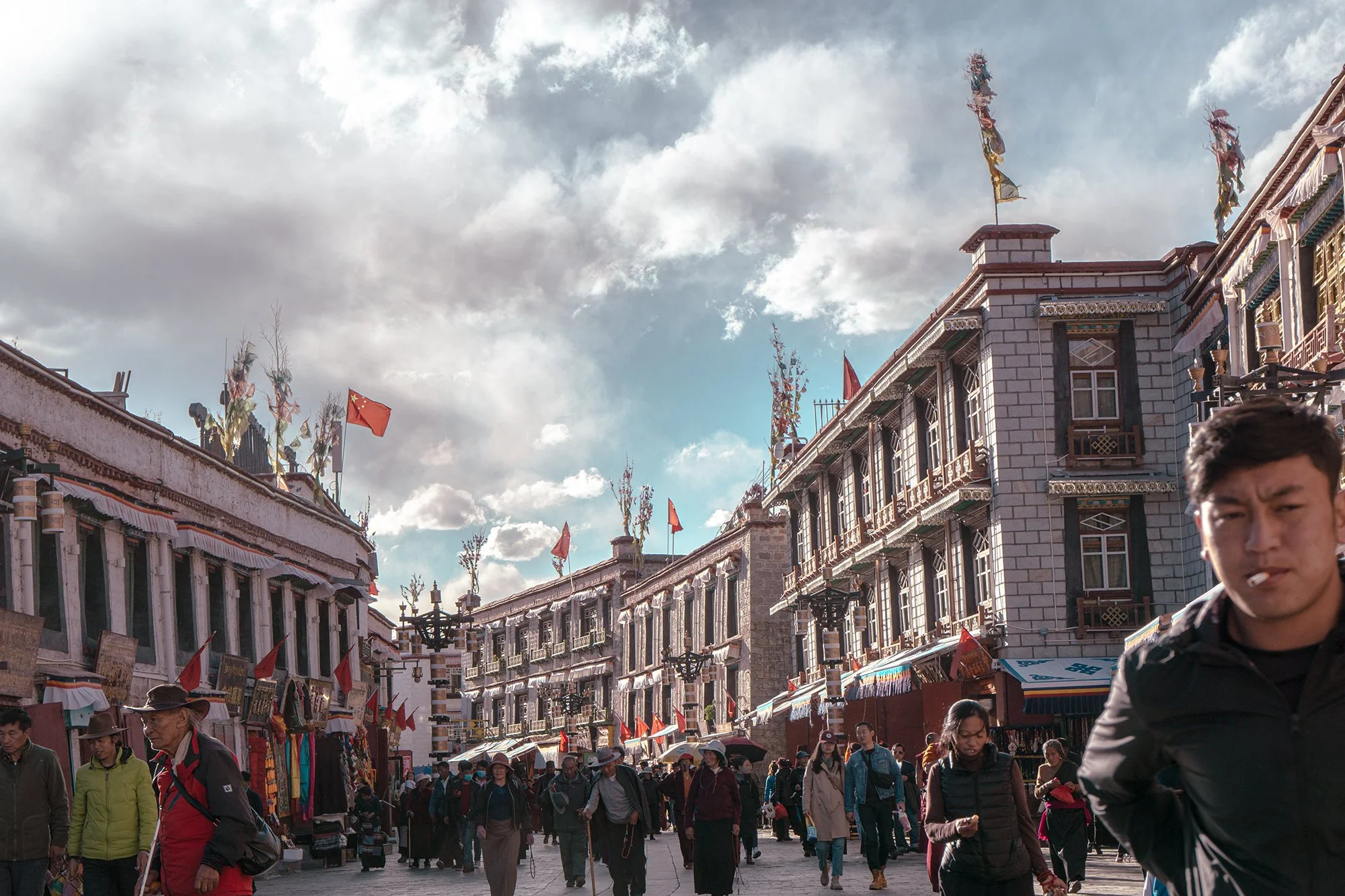 A bustling outdoor market street with many people walking, shopping, and conversing. The buildings are multi-story with ornate architecture and balconies. Flags and decorative banners are displayed on the rooftops and balconies. The sky is partly clo