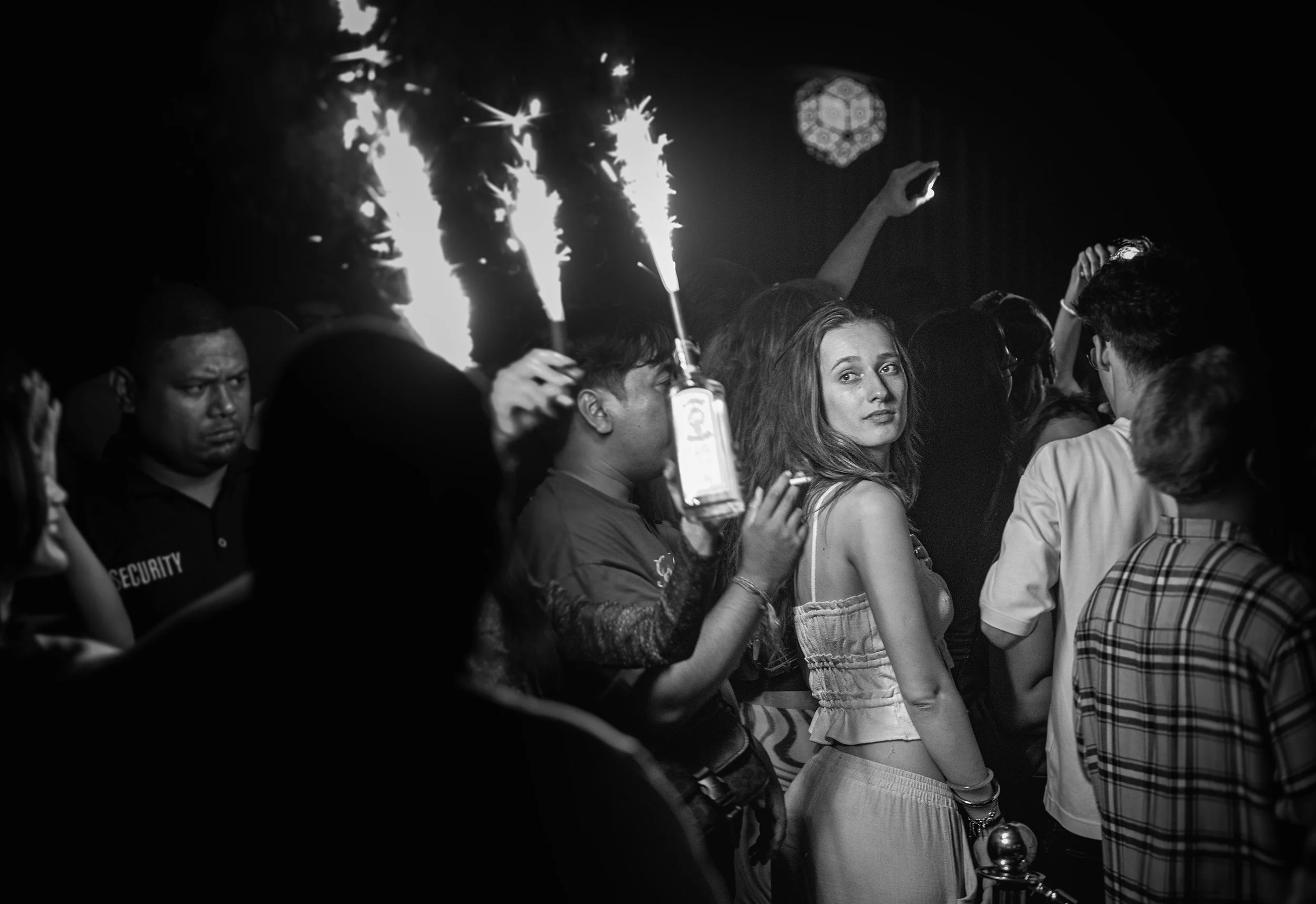 A crowd of people in a dark setting, with some holding sparklers and others taking photos or looking on. A security guard is visible on the left side of the image.
