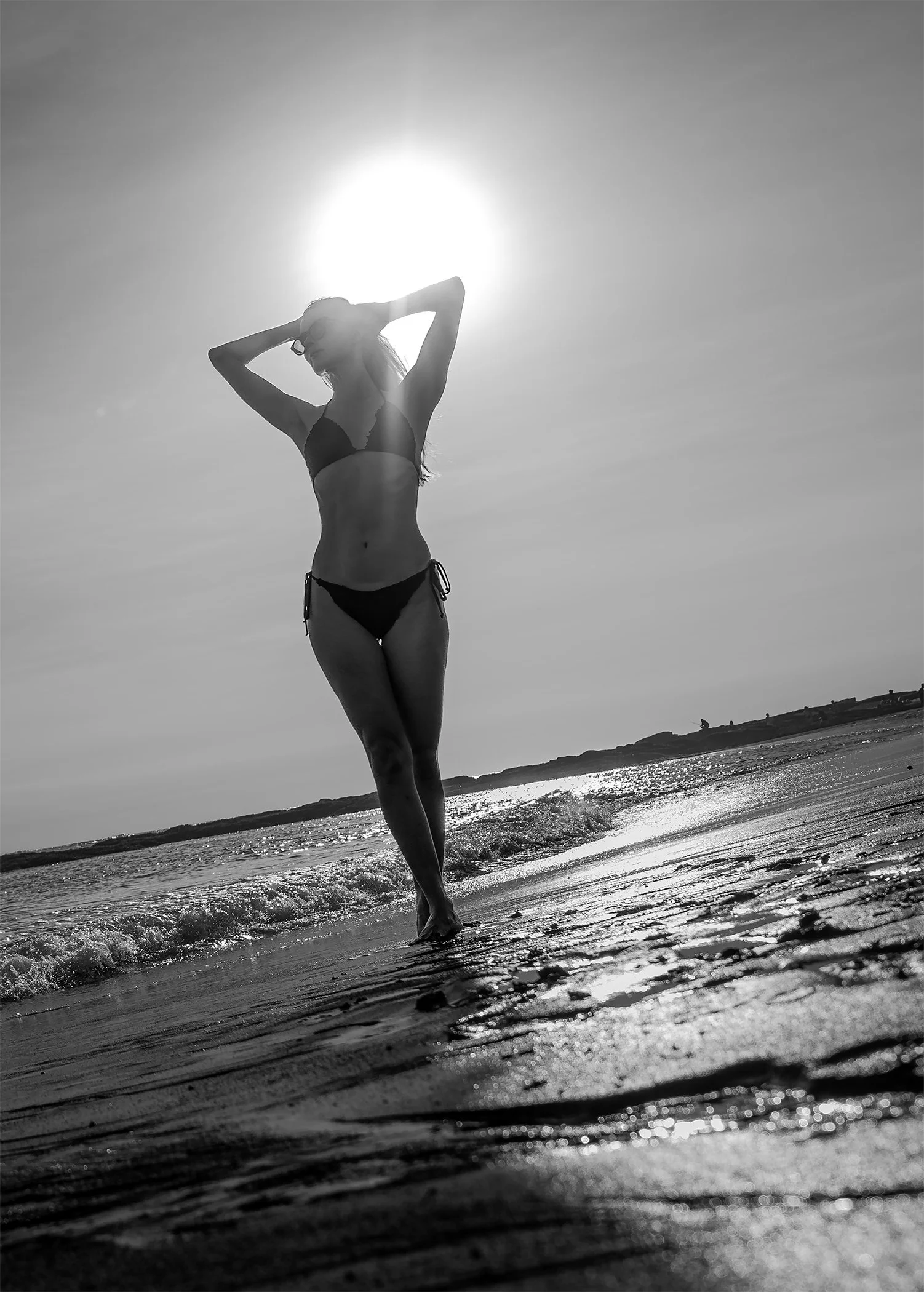 Black and white photo of a woman in a bikini walking along the beach with waves at her feet and the sun behind her.
