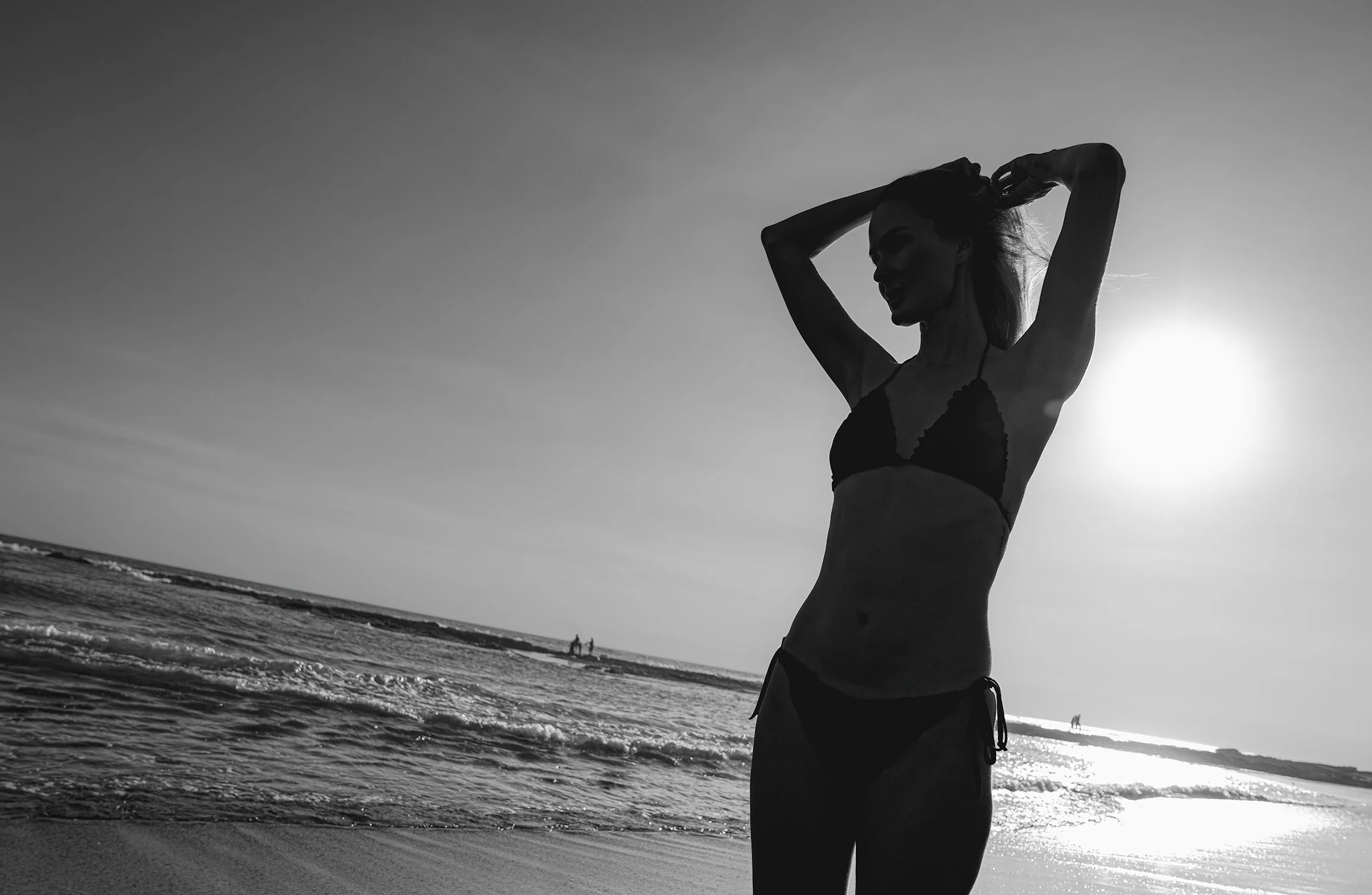 A silhouette of a woman with long hair and a bikini standing on the beach at sunset, adjusting her hair with the ocean waves in the background.
