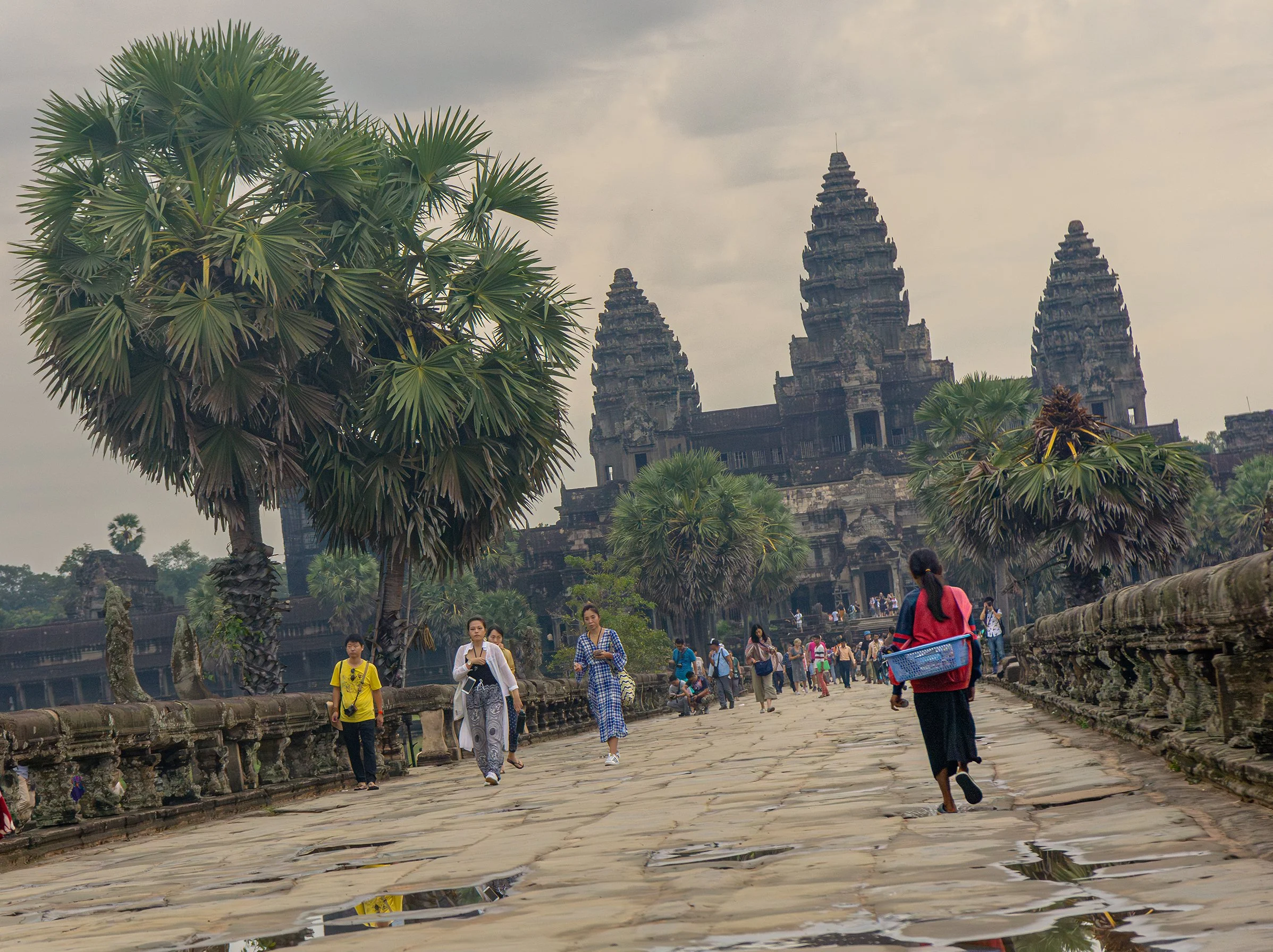 Tourists walking on a stone pathway near a historic stone temple with three tall spires, surrounded by lush greenery and large palm trees, under a cloudy sky.