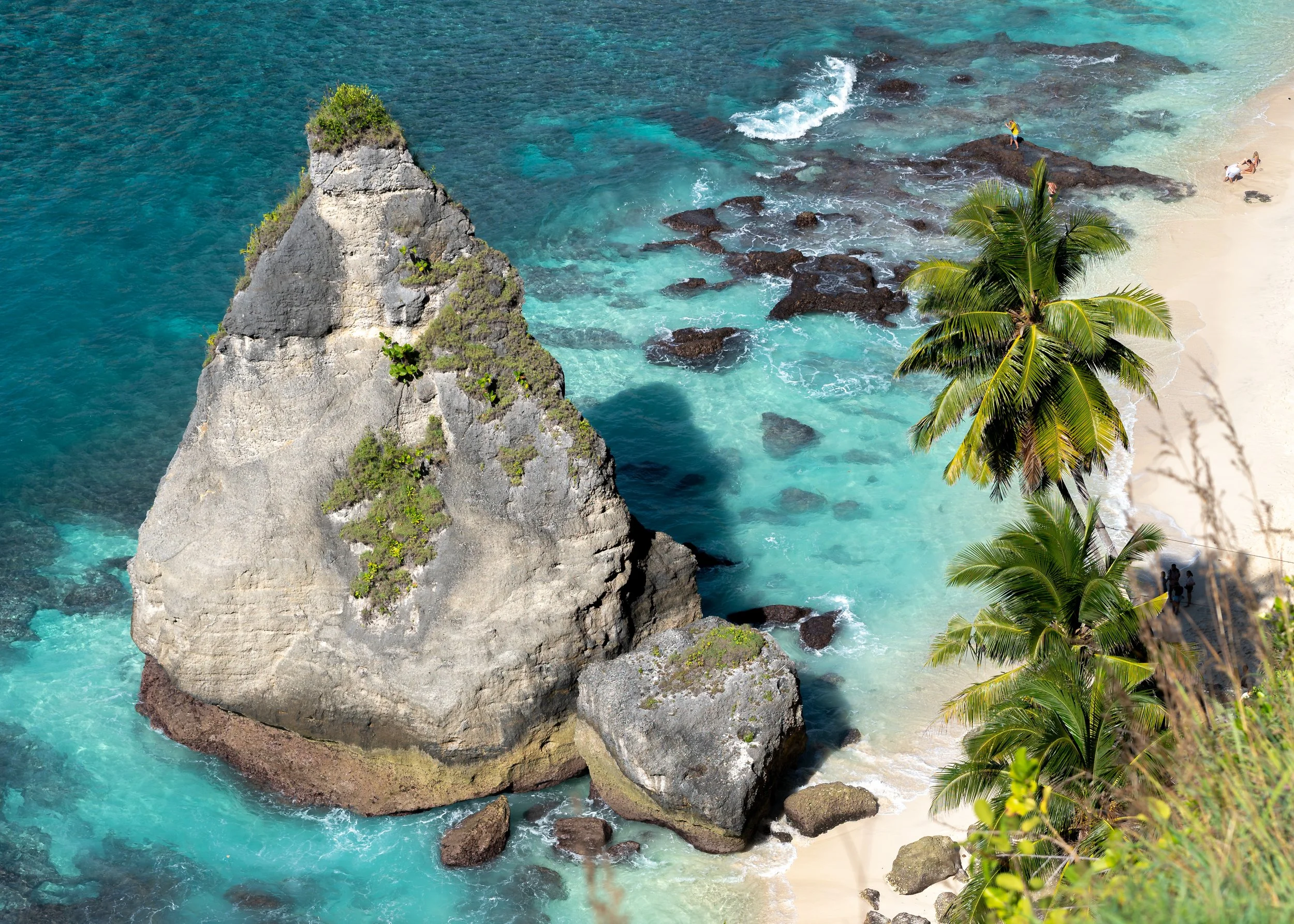 A tropical beach scene with clear turquoise water, large rocks, a small sandy beach, and palm trees.