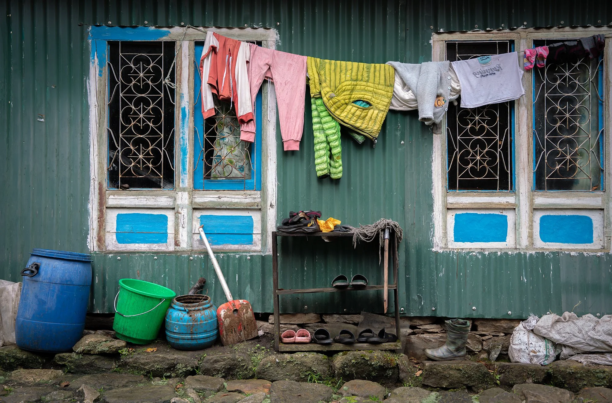 Clothes hanging on a line outside a green corrugated metal house with two windows, various boots and buckets underneath, and tools and shoes on a small outdoor shelf.