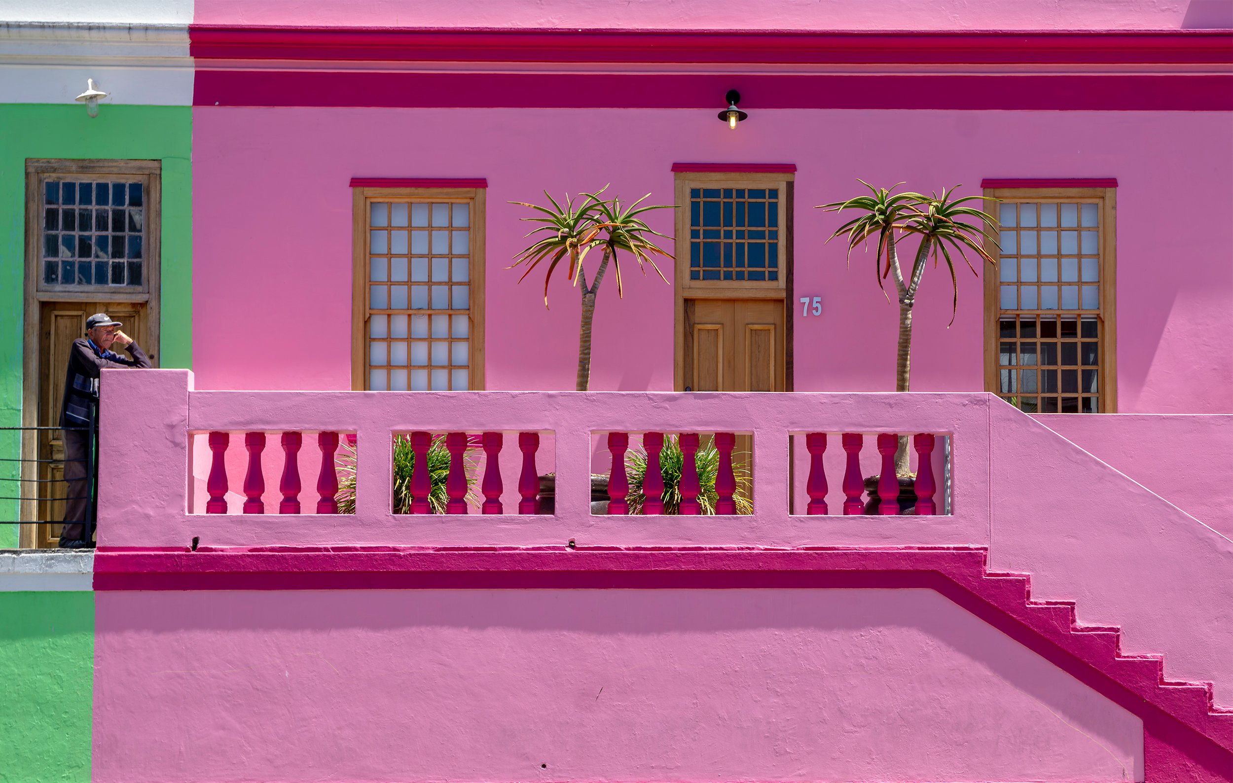 Colorful pink house with wooden windows, a pink balcony railing, and two tall tropical plants in front. There is a man standing on the left side, looking at the house.