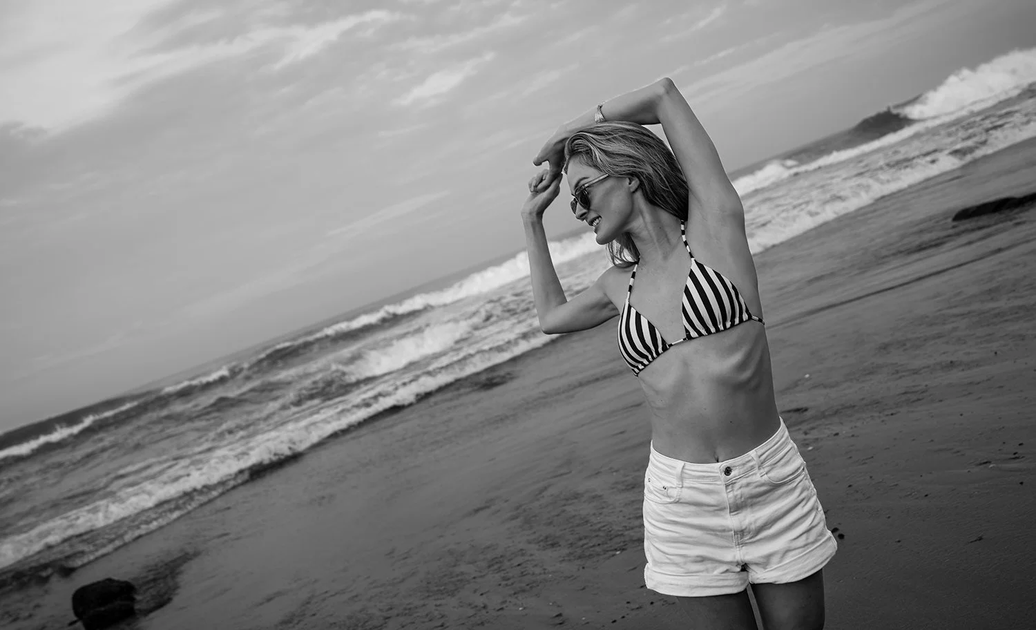 A woman in a striped bikini top and white shorts standing on the beach, smiling, with arms raised above her head, near the shoreline with waves in the background, in black and white.
