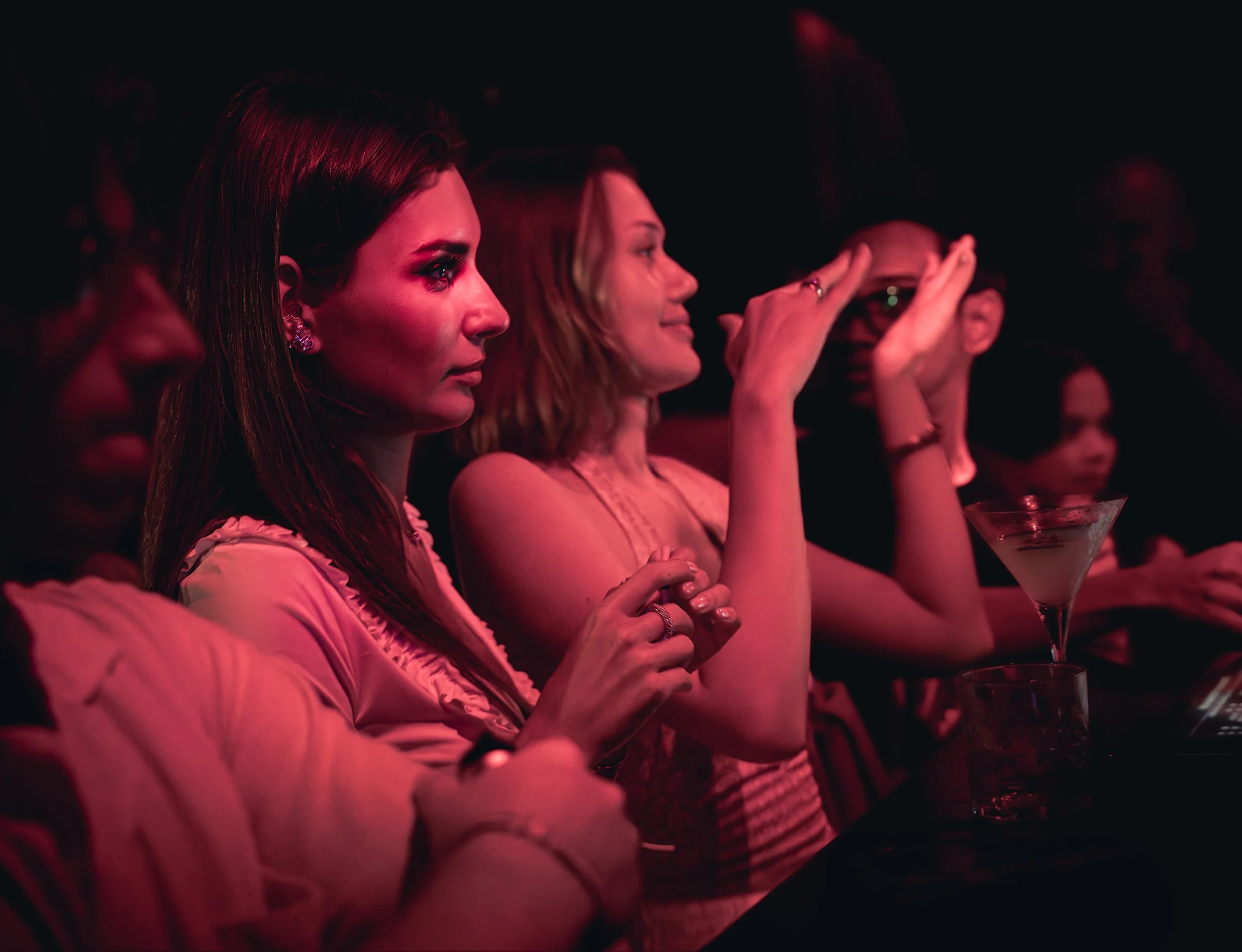 People sitting and watching an event in a dark venue, illuminated by red lighting, with one woman holding a drink in a martini glass.