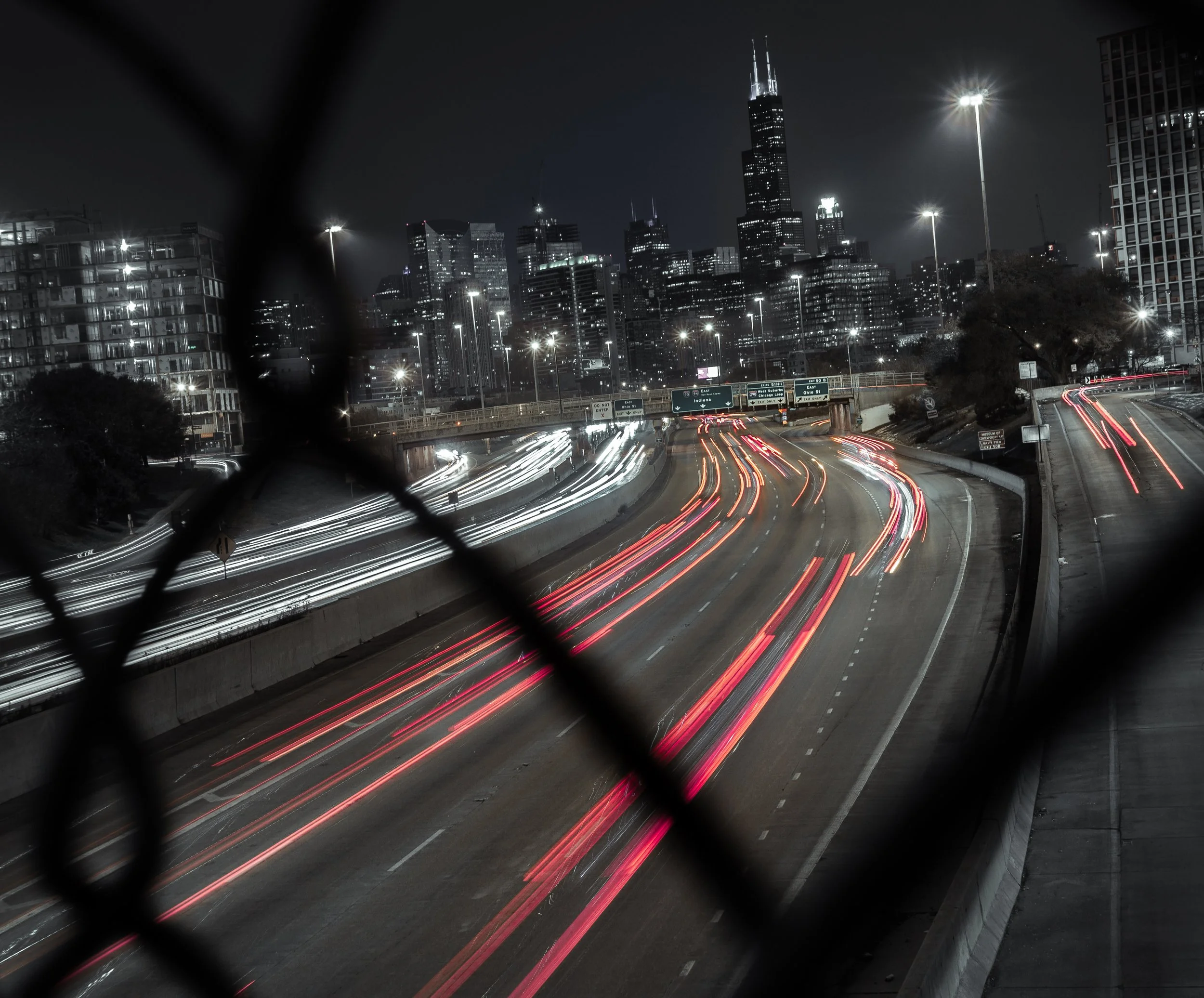 Nighttime cityscape with tall skyscrapers, including the Willis Tower, viewed through a chain-link fence. Light trails from moving vehicles on a highway are visible in the foreground.