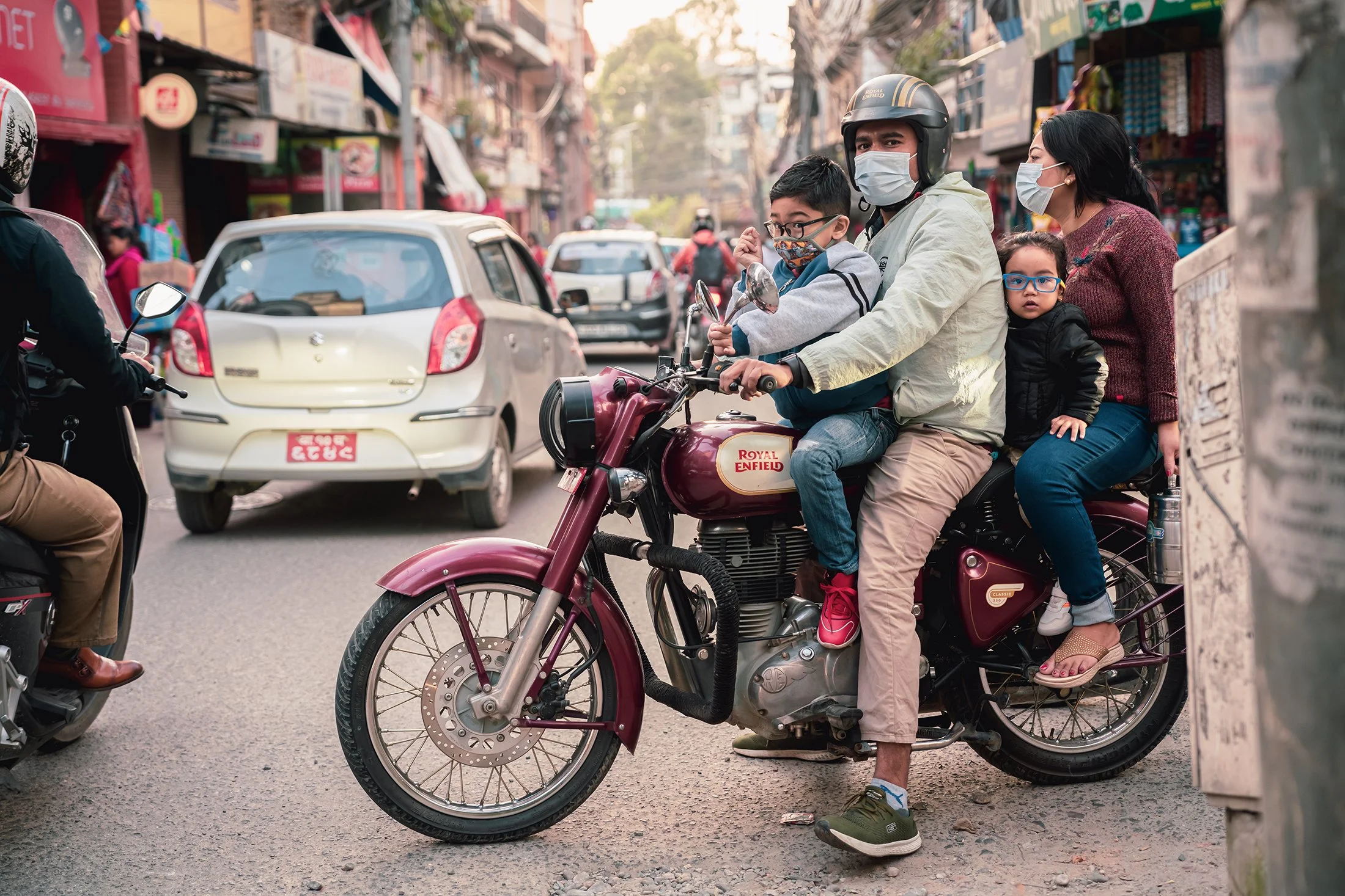 A group of five people, two children and three adults, sitting on a maroon Royal Enfield motorcycle in a busy street with cars and shops. They are all wearing face masks.