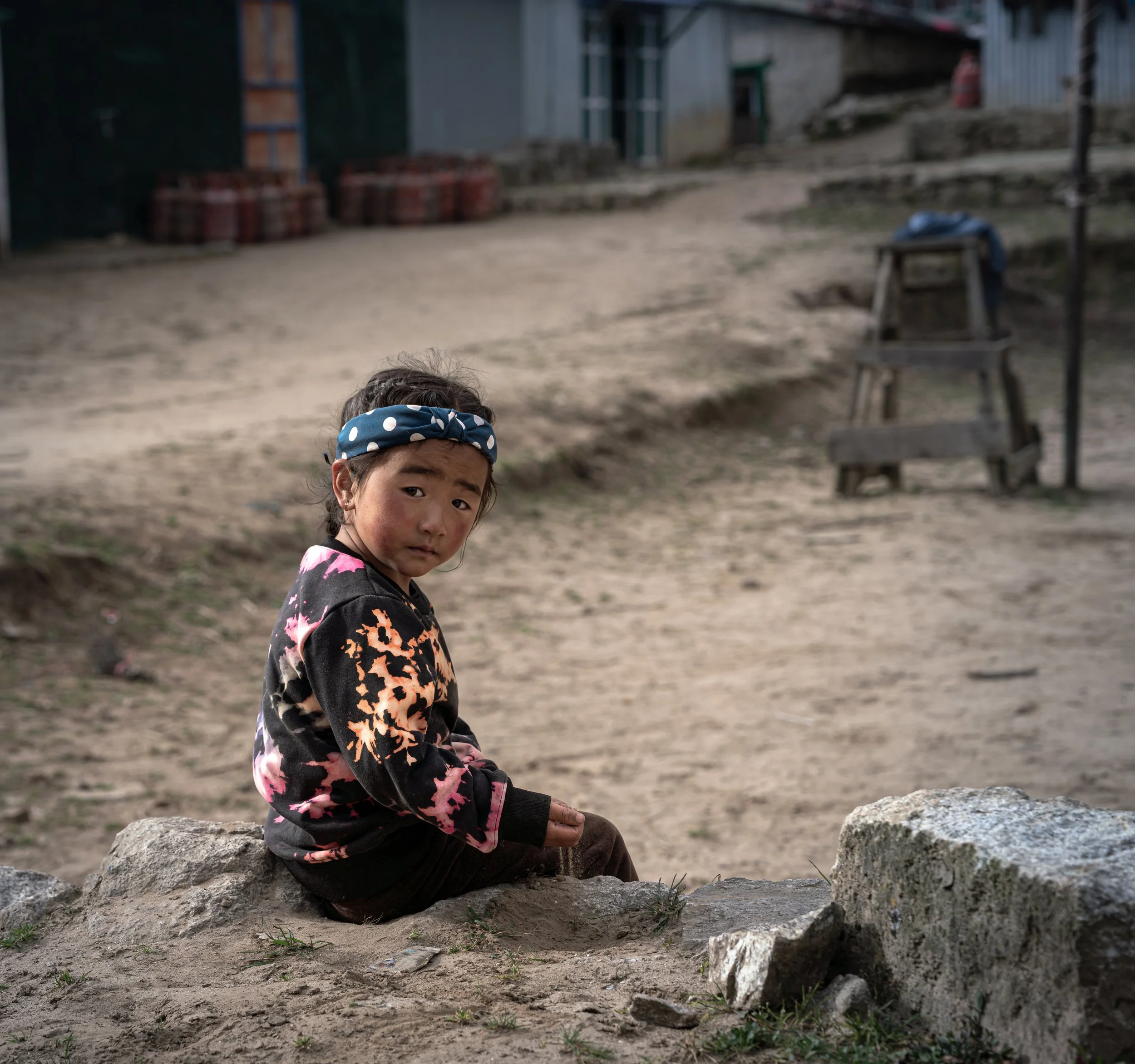 A young girl with a blue polka dot headband sitting on rocks in an outdoor setting with buildings and stairs in the background.