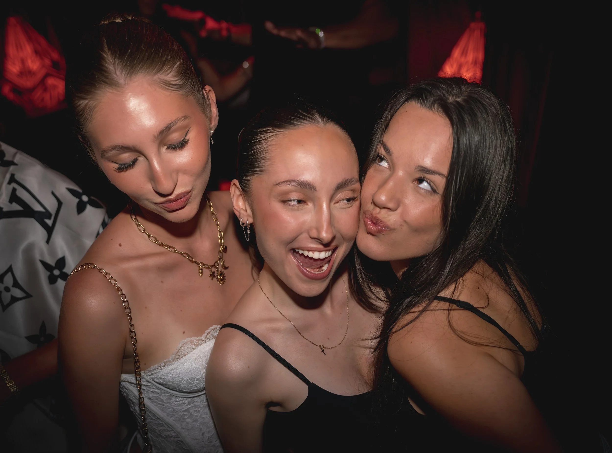 Four women at a social event taking a close-up selfie together, with affectionate expressions and makeup, against a dimly lit background.