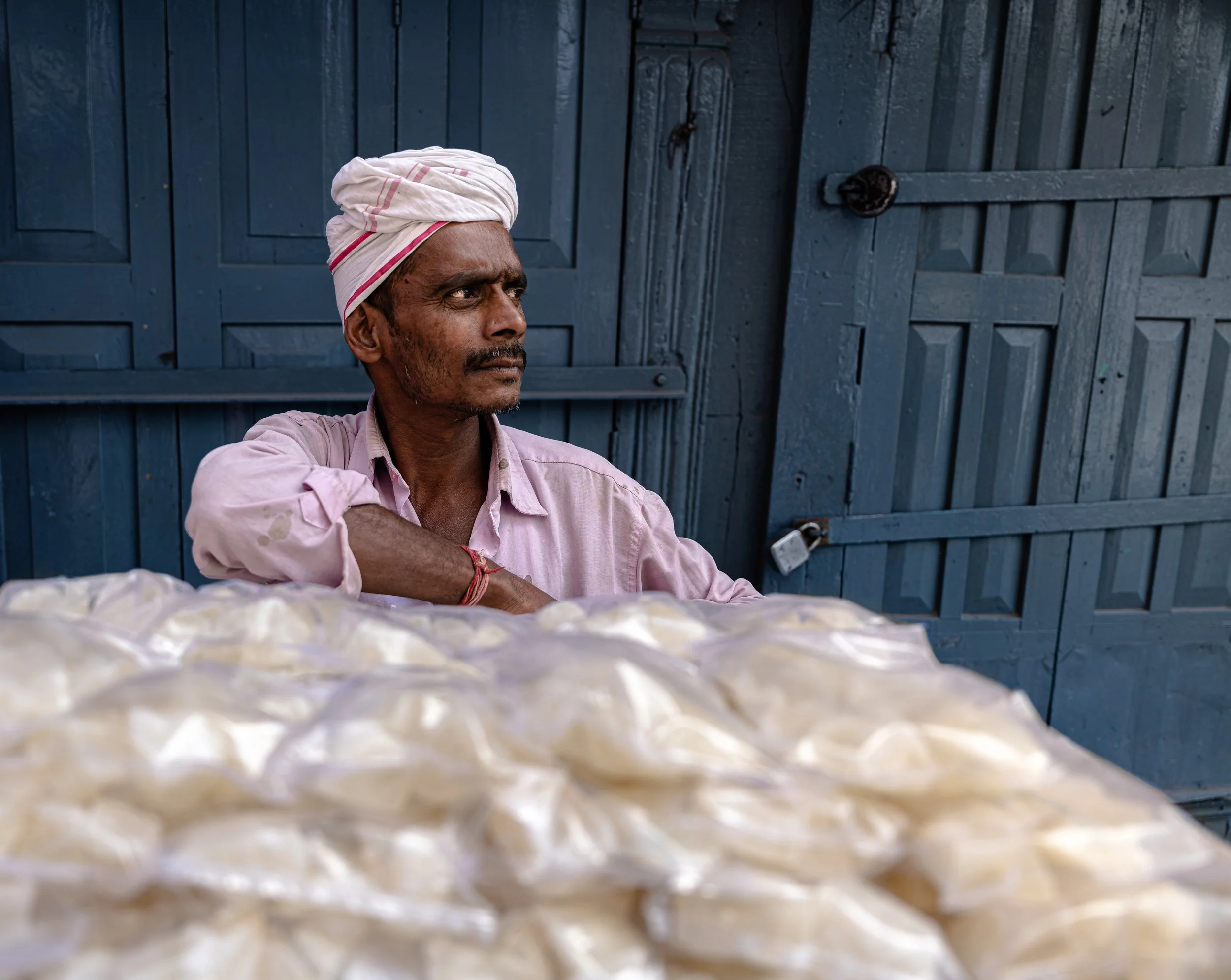 A man wearing a pink shirt and a white turban sits in front of a blue wooden door.