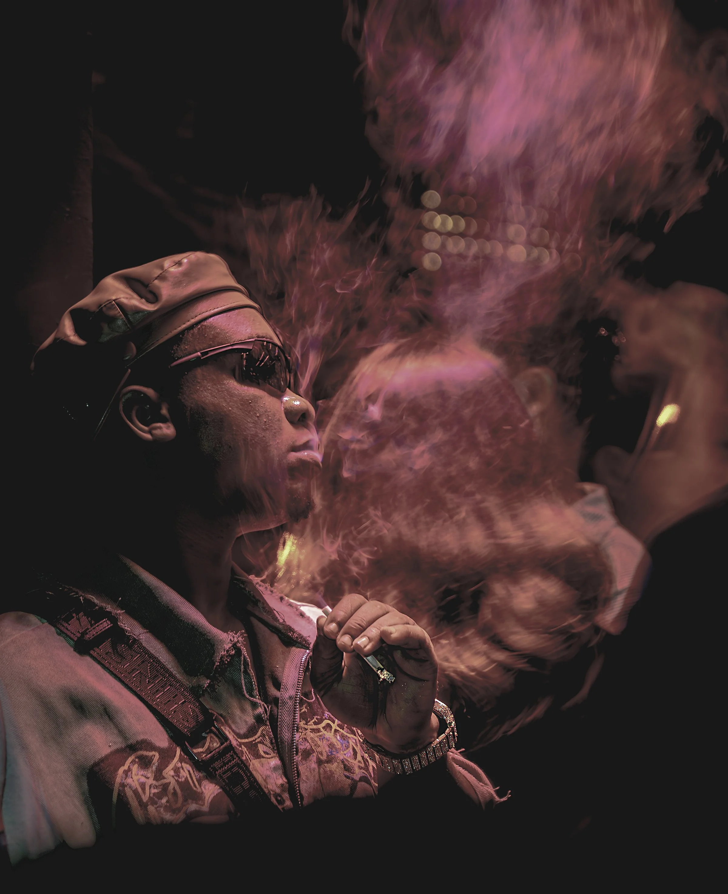 A person with glasses and a cap, illuminated by pink and purple light, smoking a vape or e-cigarette with visible vapor around their face.