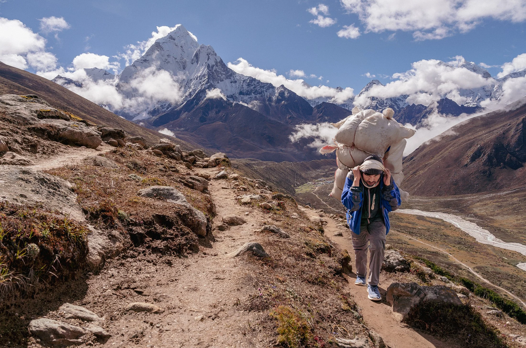 A man hiking on a rugged trail in a mountainous landscape with snow-capped peaks and clouds in the background, carrying a large load on his back.