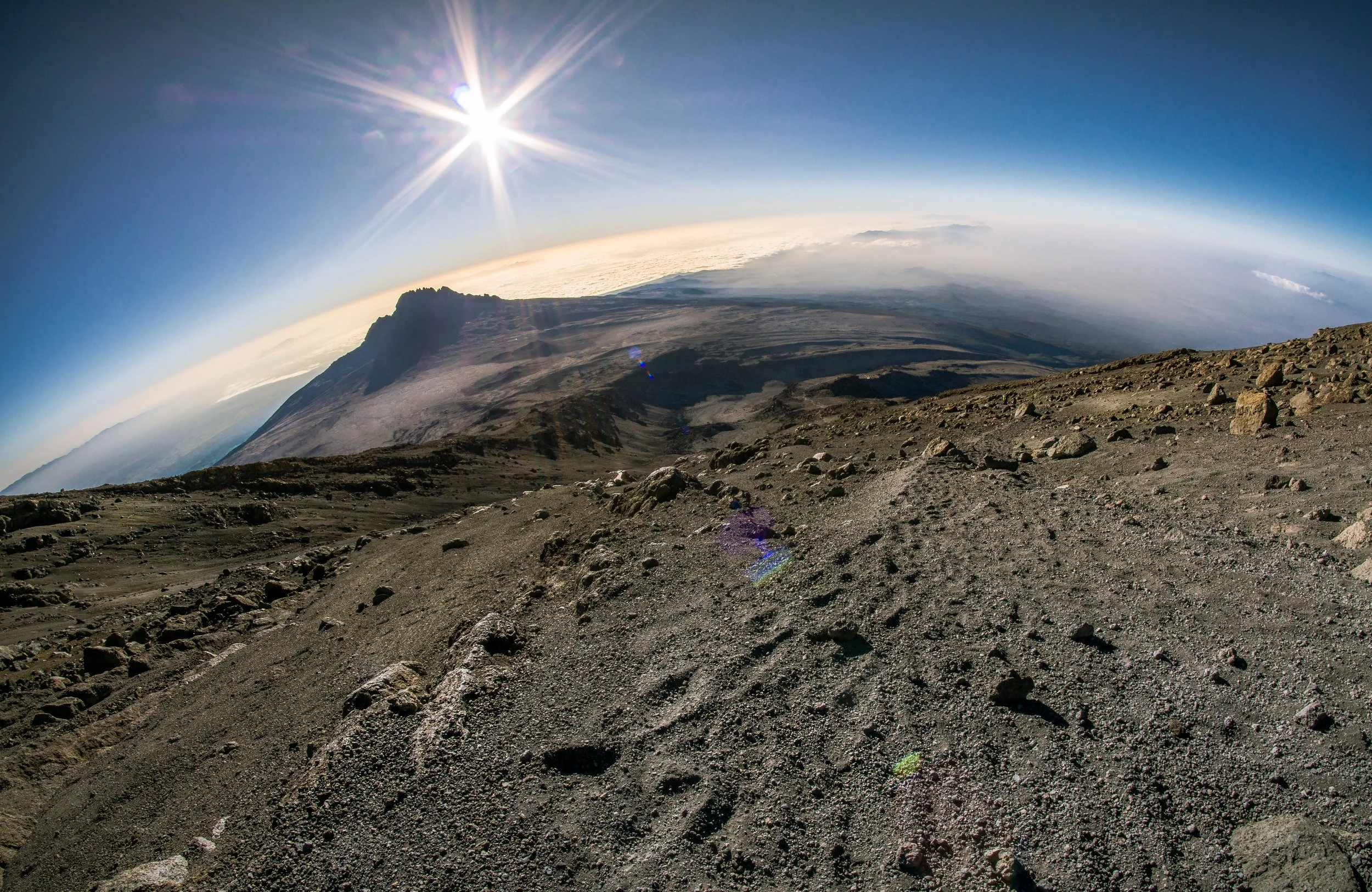 View from the top of a volcanic mountain showing rocky terrain, a distant mountain peak, clouds, and the sun shining brightly in a clear blue sky.