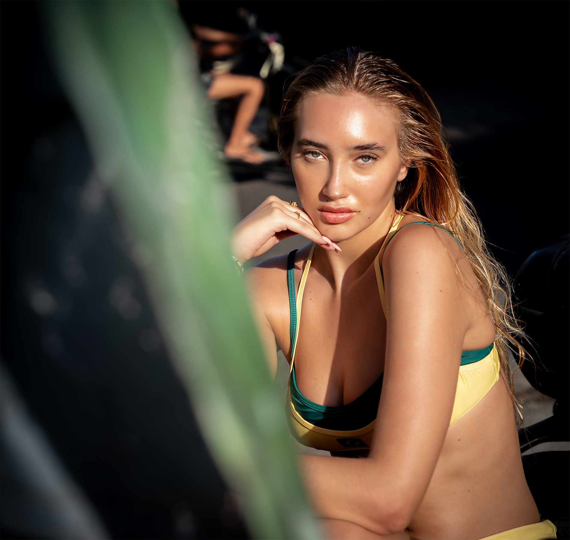 A woman with wet hair wearing a yellow and teal sports bra looking at the camera, with a blurred person in the background