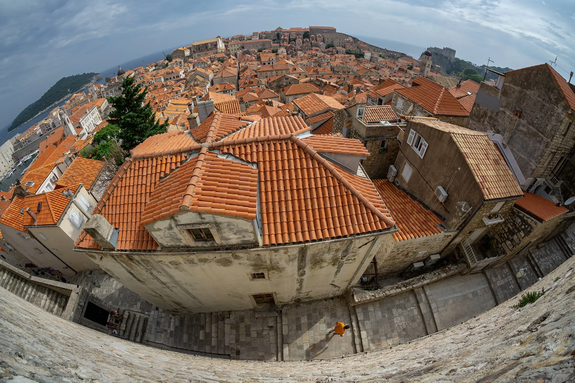 A panoramic view of a historic city with orange-tiled rooftops on a hillside, overlooking the sea, with cloudy skies above.