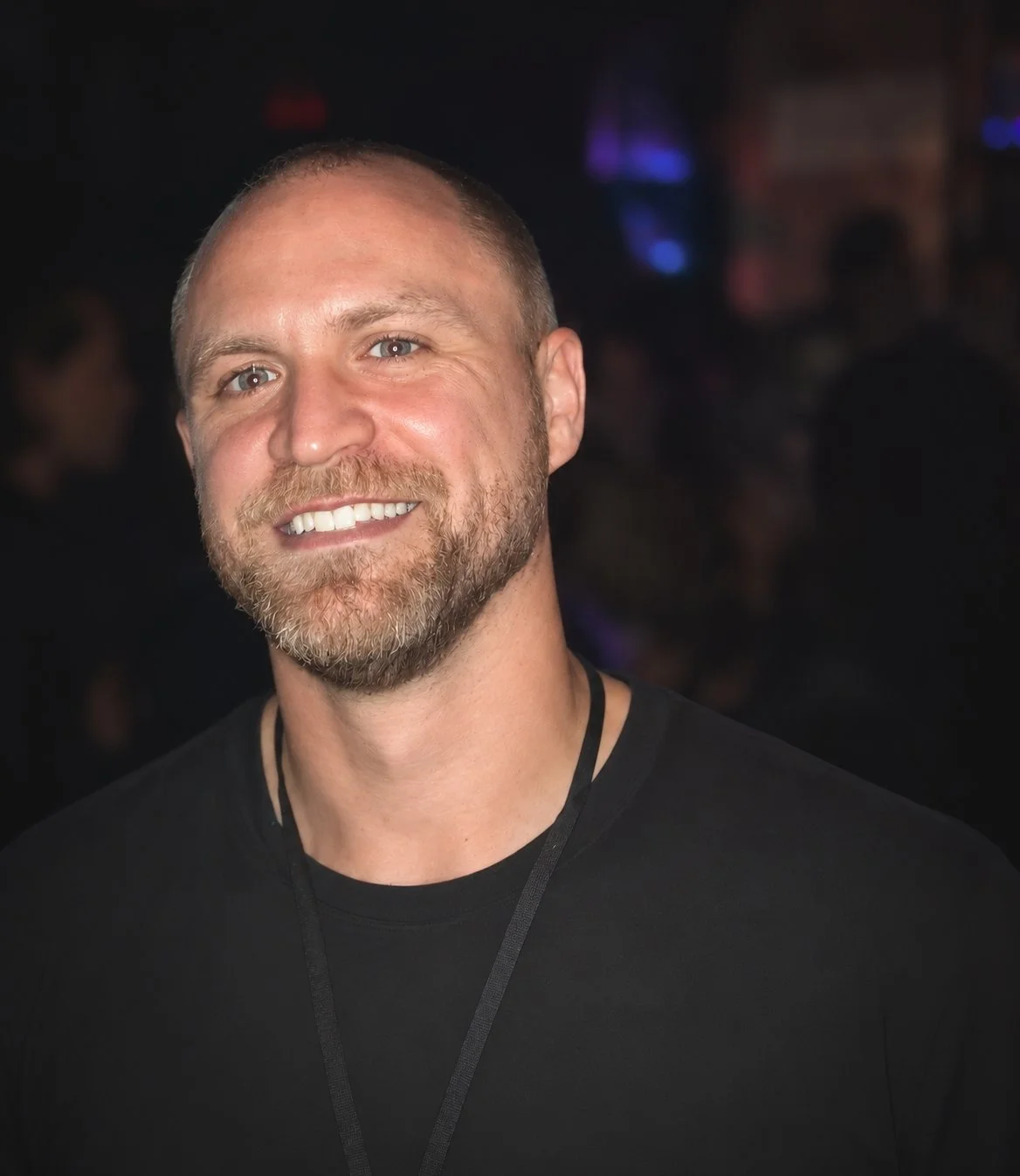 A smiling man with a beard and short hair, wearing a black shirt, in a dimly lit environment.