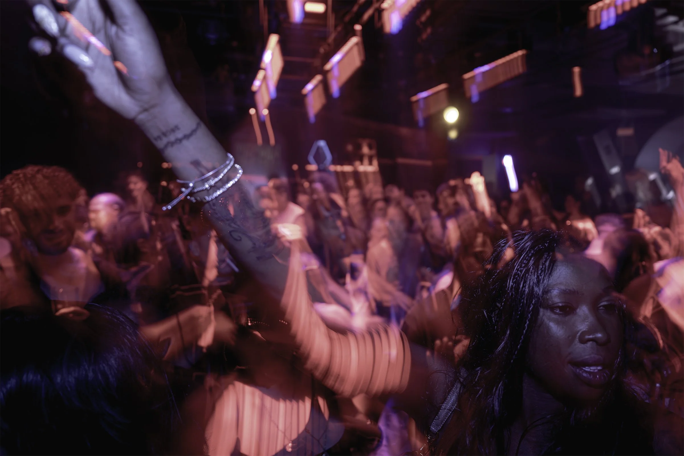 A crowded dance floor at a nightclub with pink and purple neon lighting, a person with tattoos and jewelry raising their arm, and others enjoying the music.