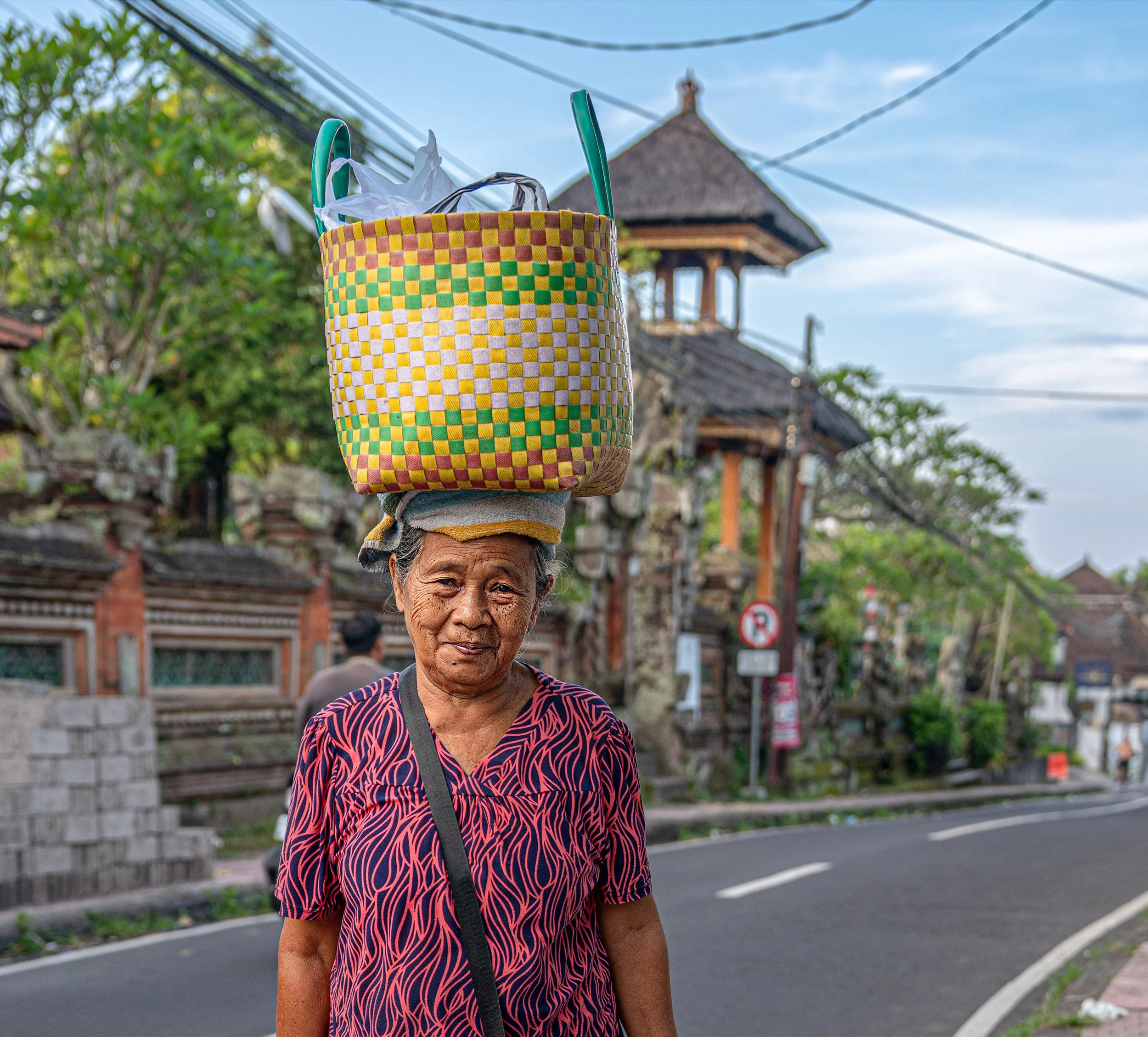 An elderly woman with a checkered red, yellow, green, and white basket balanced on her head, wearing a dark top with red patterns and a black strap across her shoulder, standing outdoors near a street with trees and buildings in the background.