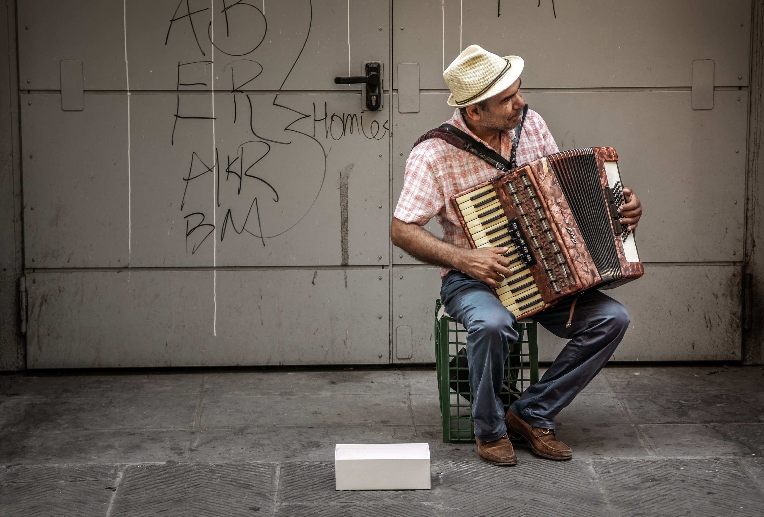 A man wearing a beige hat and red checkered shirt sits on a small green stool, playing an accordion. He is positioned against a gray metal door with graffiti and graffiti tags in black marker.