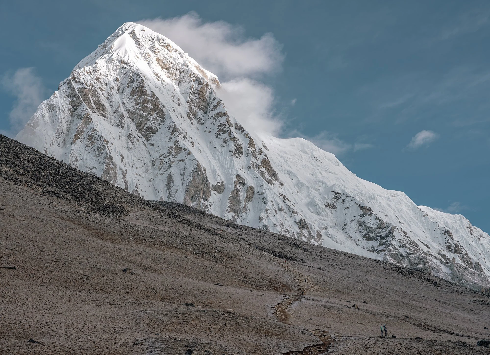Snow-covered mountain with rugged slopes under a partly cloudy sky, with small figures of hikers on a gravel path at the base.