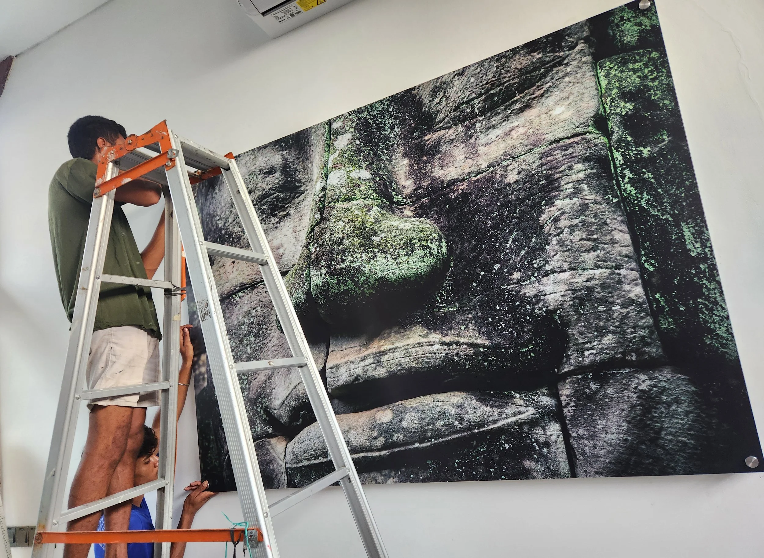 Several people are setting up or installing a large artwork on a white wall. The artwork depicts a rocky landscape with moss, and the scene appears to be a large photo or mural. A person on a ladder is working on the top part, while others are assisting from below.