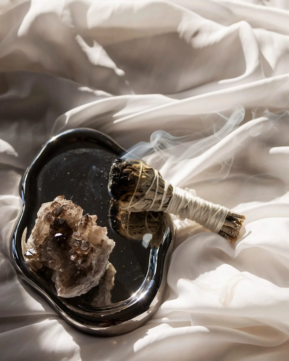A ritual sage bundle and raw crystal on a silver tray with flowing white linen, representing the Quiet pillar of the Suma wellness residency.