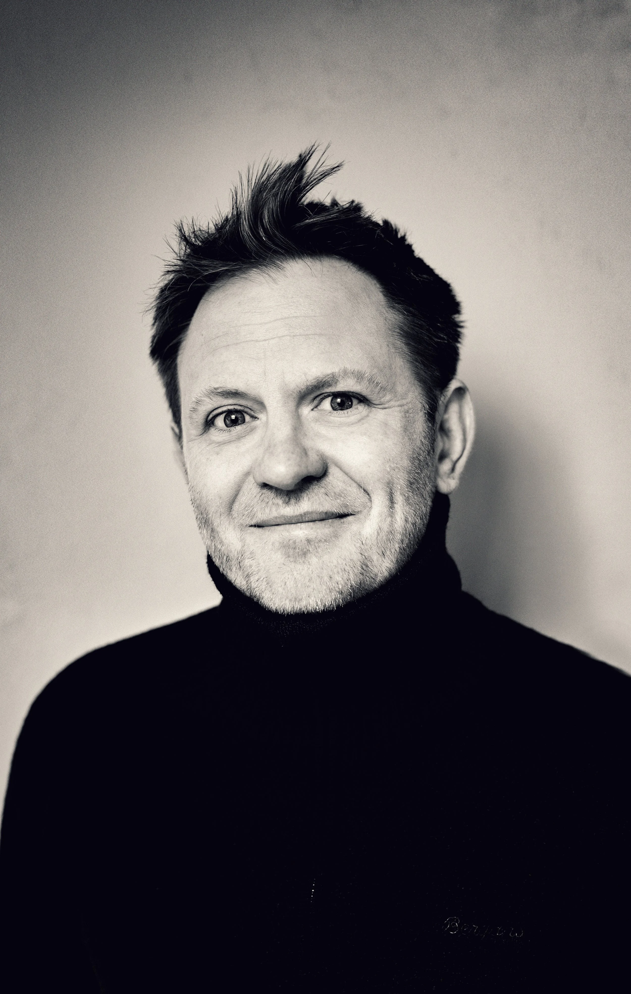 Sort-hvid portrætfotografi af en mand med kort, opskåret hår, iført sort turtleneck, smilende. Billedet er taget i studiemiljø med en ensfarvet baggrund.