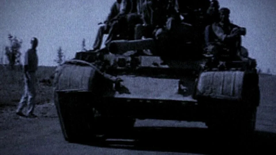 En sort tankskrog med soldater, der kører på en åben mark, med to personer i baggrunden.