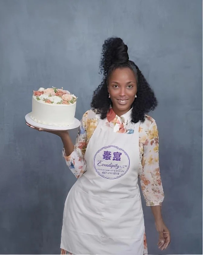 Woman holding a decorated birthday cake, wearing a floral blouse and apron, standing against a plain gray background.