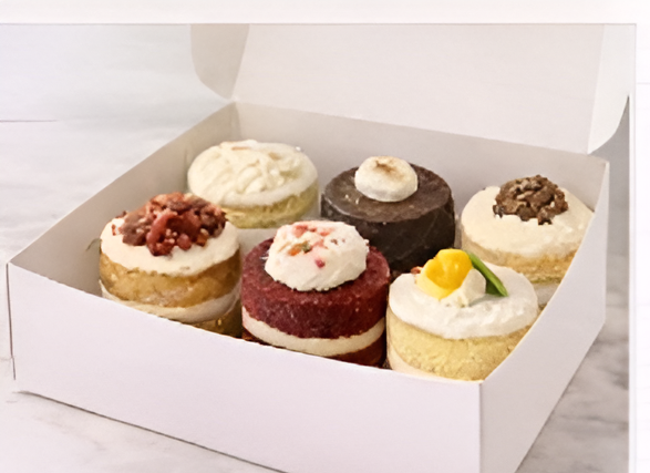 Six cupcakes decorated with various toppings in a white bakery box.
