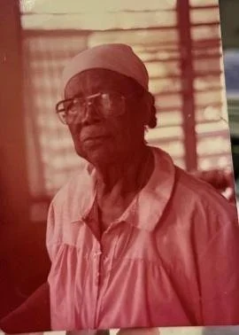 An elderly woman wearing glasses, a light-colored headscarf, and a matching shirt sitting indoors with a window and wooden blinds in the background.