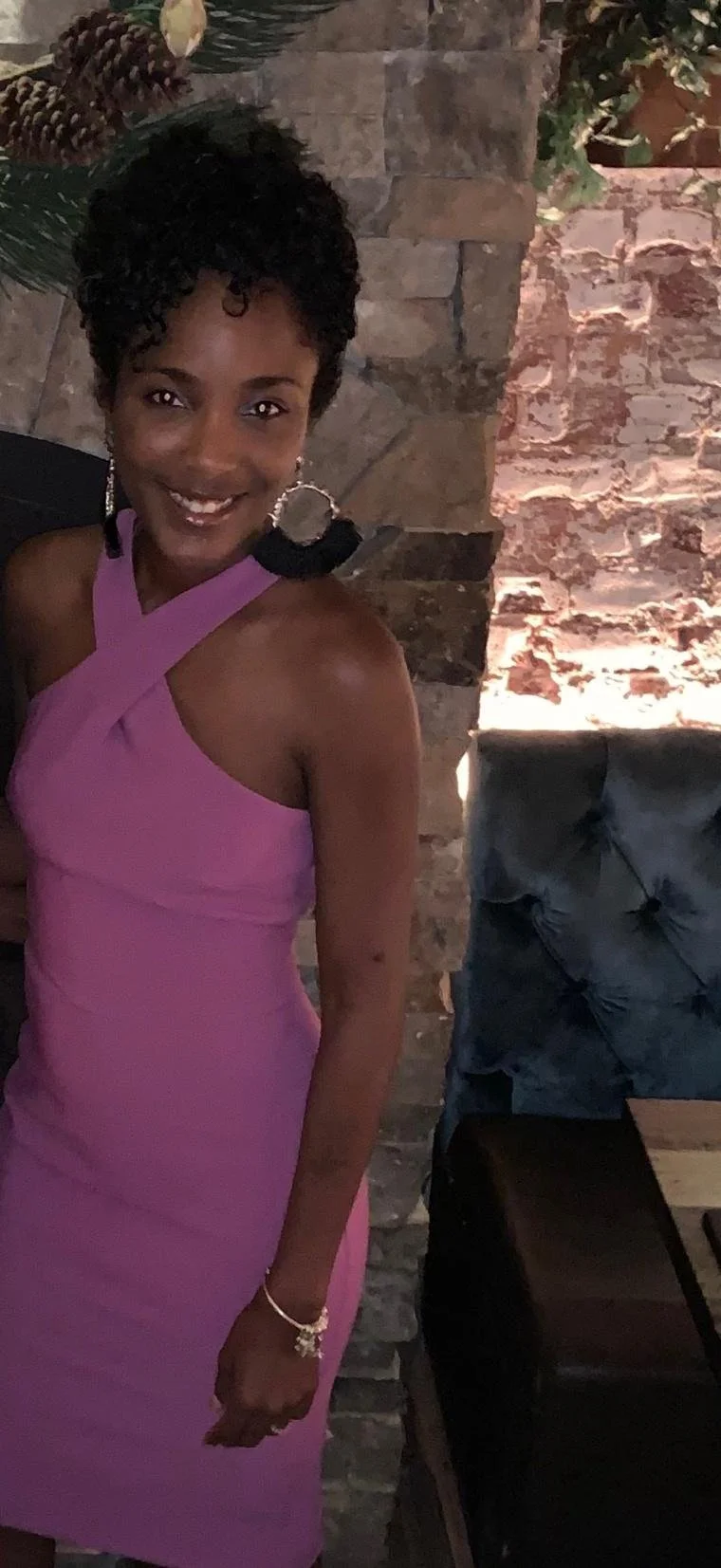 A woman smiling, wearing a pink dress with an asymmetrical strap design, standing against a stone wall with a cushioned chair nearby.