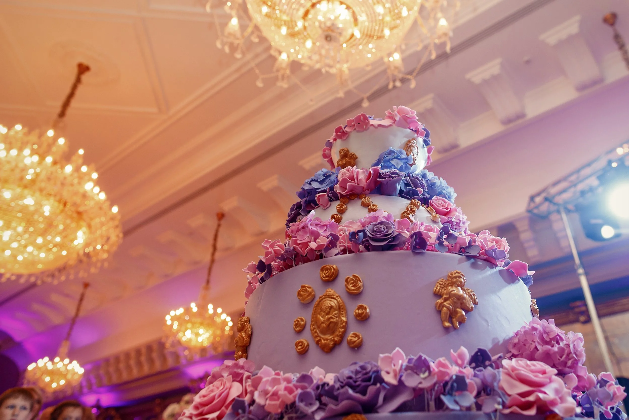 A multi-tiered wedding cake decorated with purple, pink, and blue flowers and gold embellishments, set in an elegant ballroom with chandeliers and pink lighting.